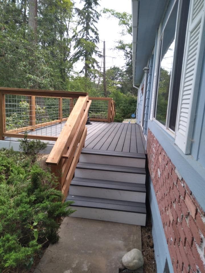 A wooden deck with a staircase leads up to a house with blue siding and brick accents beside a paved walkway and trees.