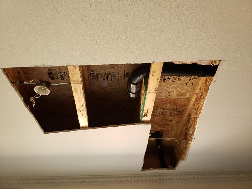 A rectangular opening in a residential ceiling showing exposed wooden joists, electrical wiring, and black plumbing pipes.