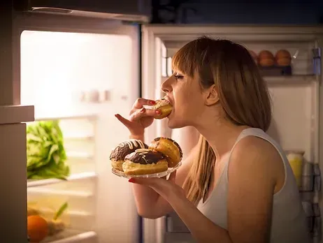 A woman is eating donuts in front of an open refrigerator.