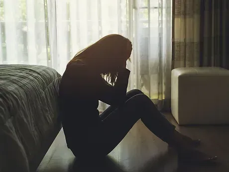 A woman is sitting on the floor in front of a bed with her head in her hands.