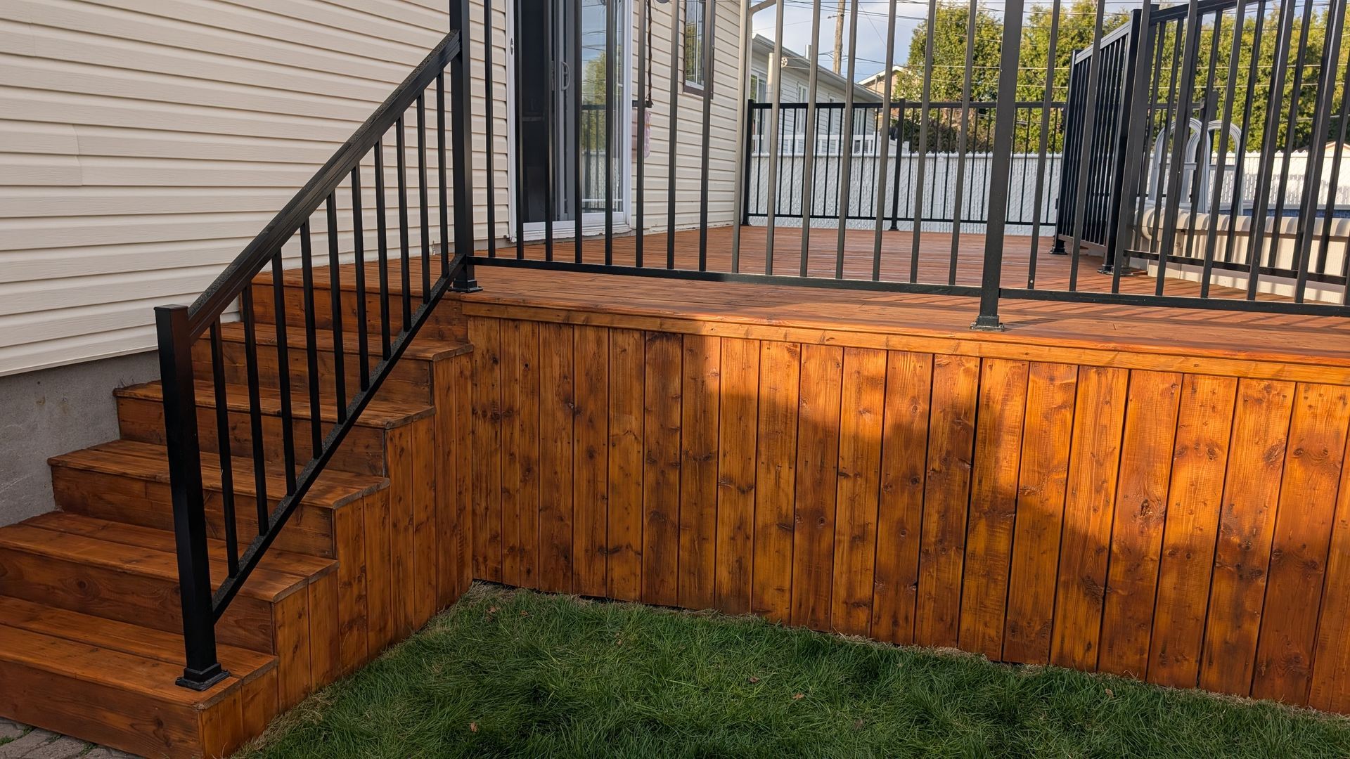 Terrasse en bois avec rambarde et marches noires, adossée à un bâtiment de couleur claire, avec de l'herbe verte au premier plan.