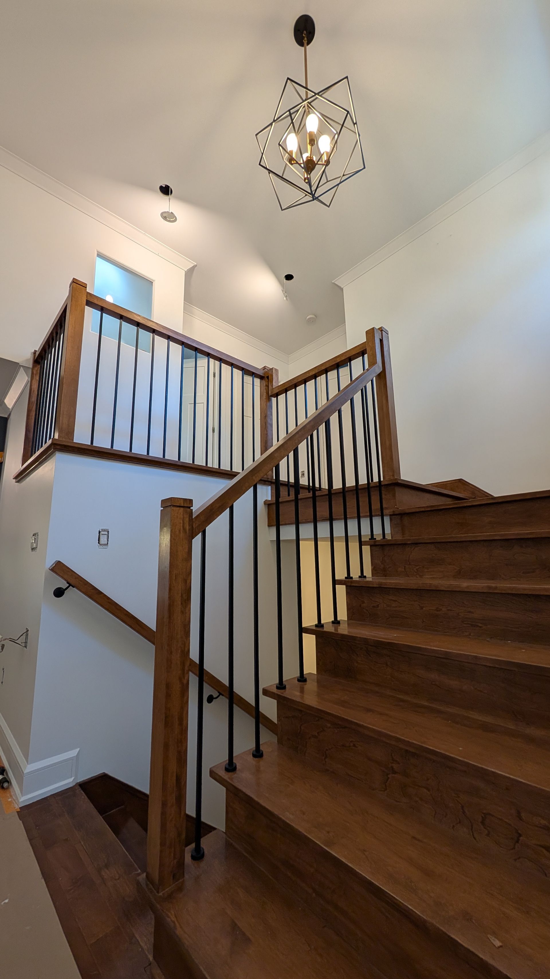 Escalier en bois avec rampes noires, menant au deuxième étage. Lustre géométrique et éclairage encastré.