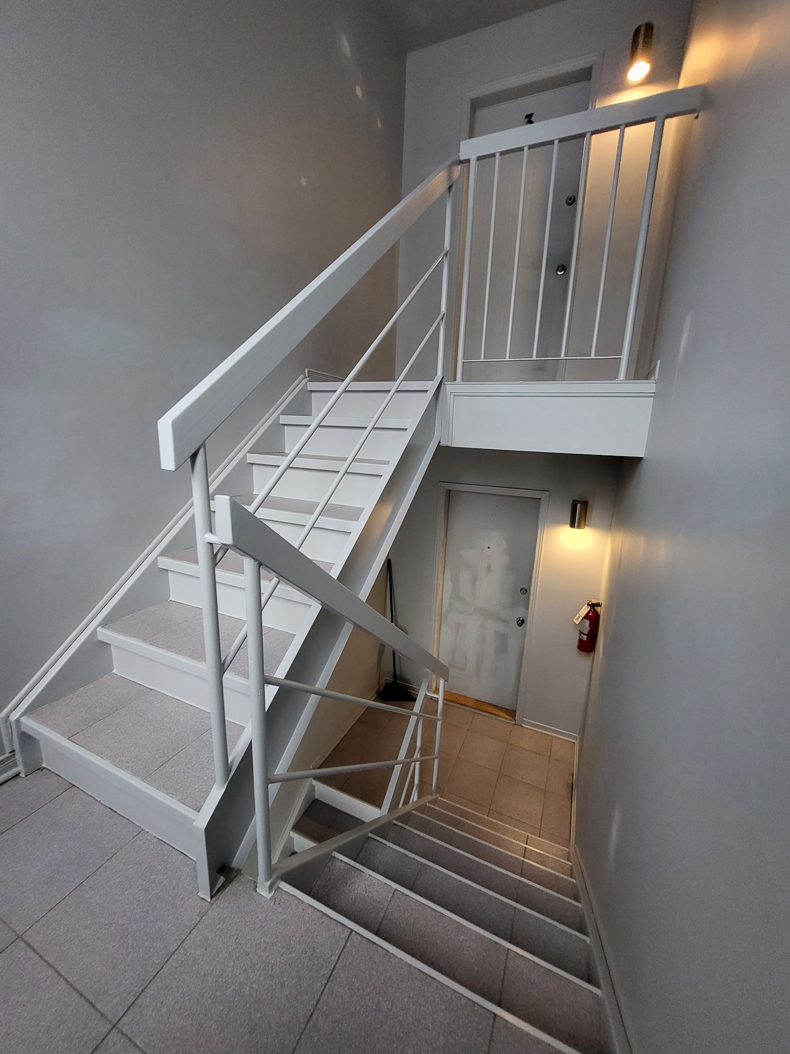 Escalier aux murs gris et à la rampe blanche. Un extincteur est visible.