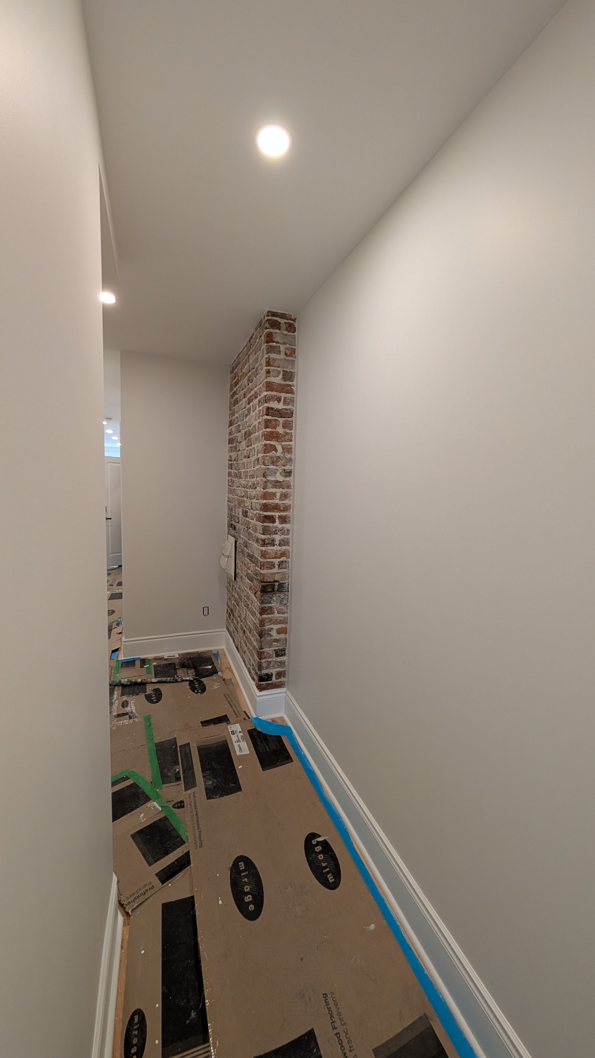Couloir étroit avec une section de mur en briques apparentes, des murs gris et un revêtement de sol protecteur.