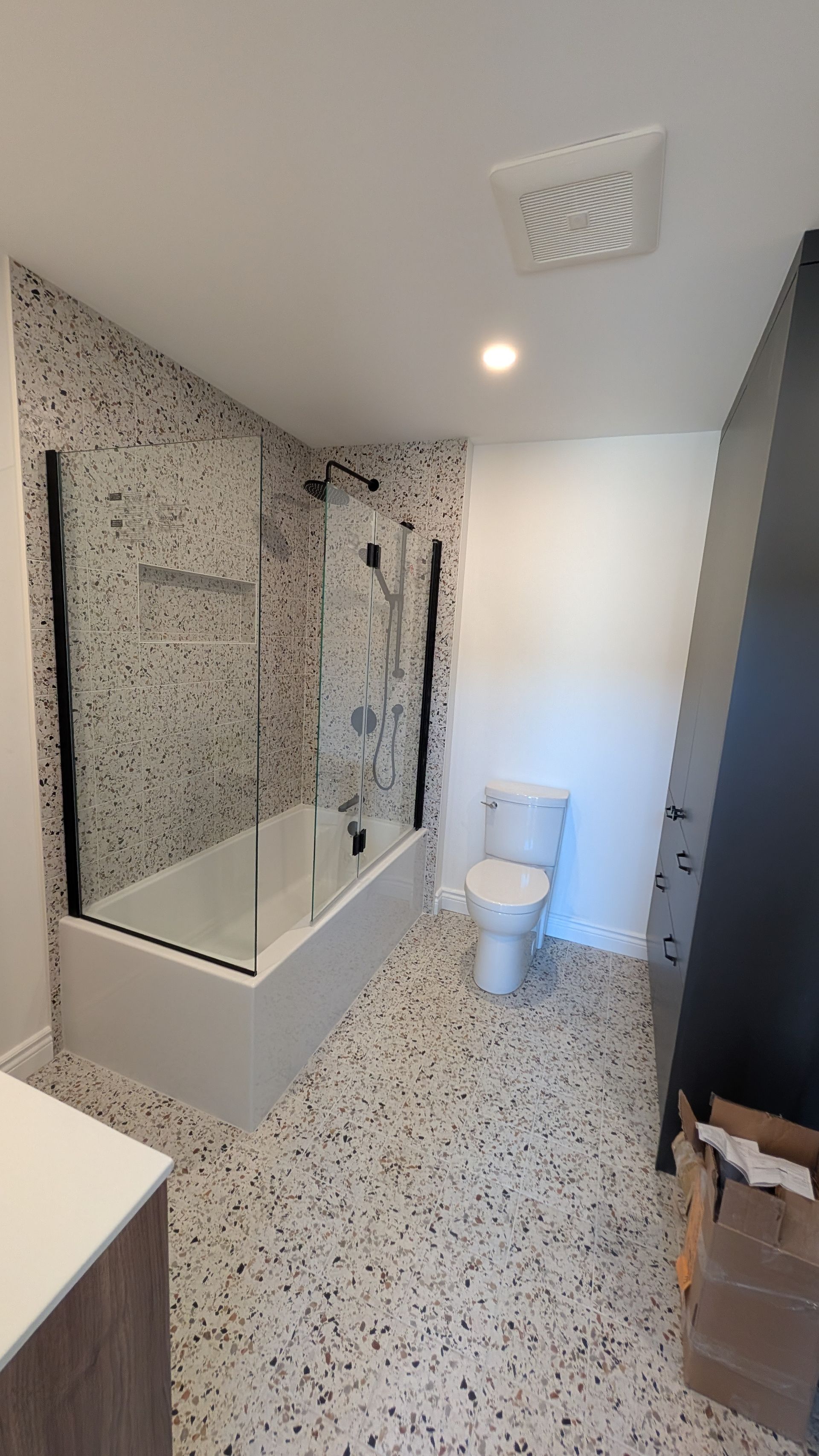 Salle de bains moderne avec baignoire et douche, mur en mosaïque, sol en terrazzo, toilettes blanches et cadre de douche noir.