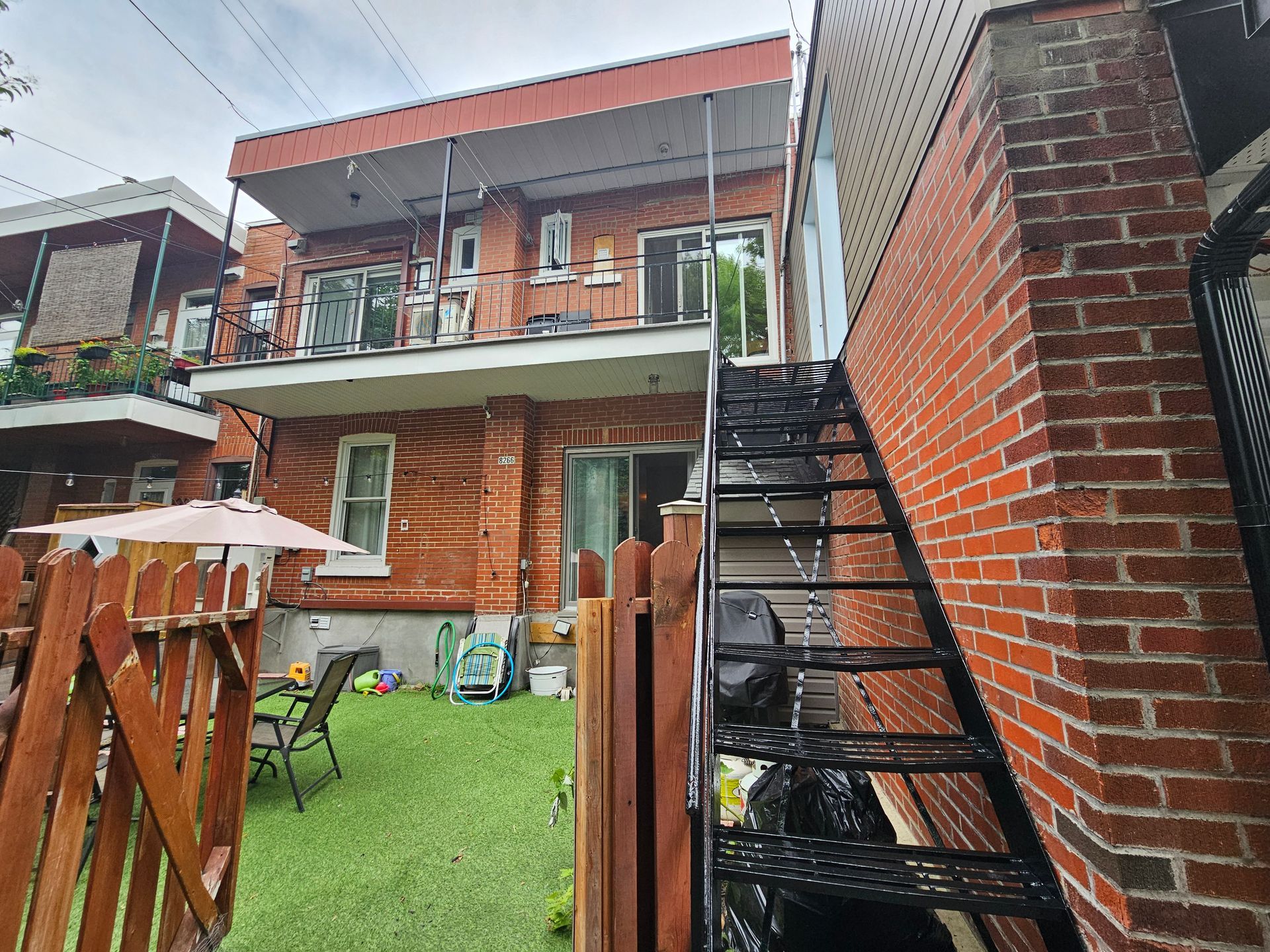 Vue arrière d'une maison en briques avec un escalier extérieur en métal et une petite clôture en bois.