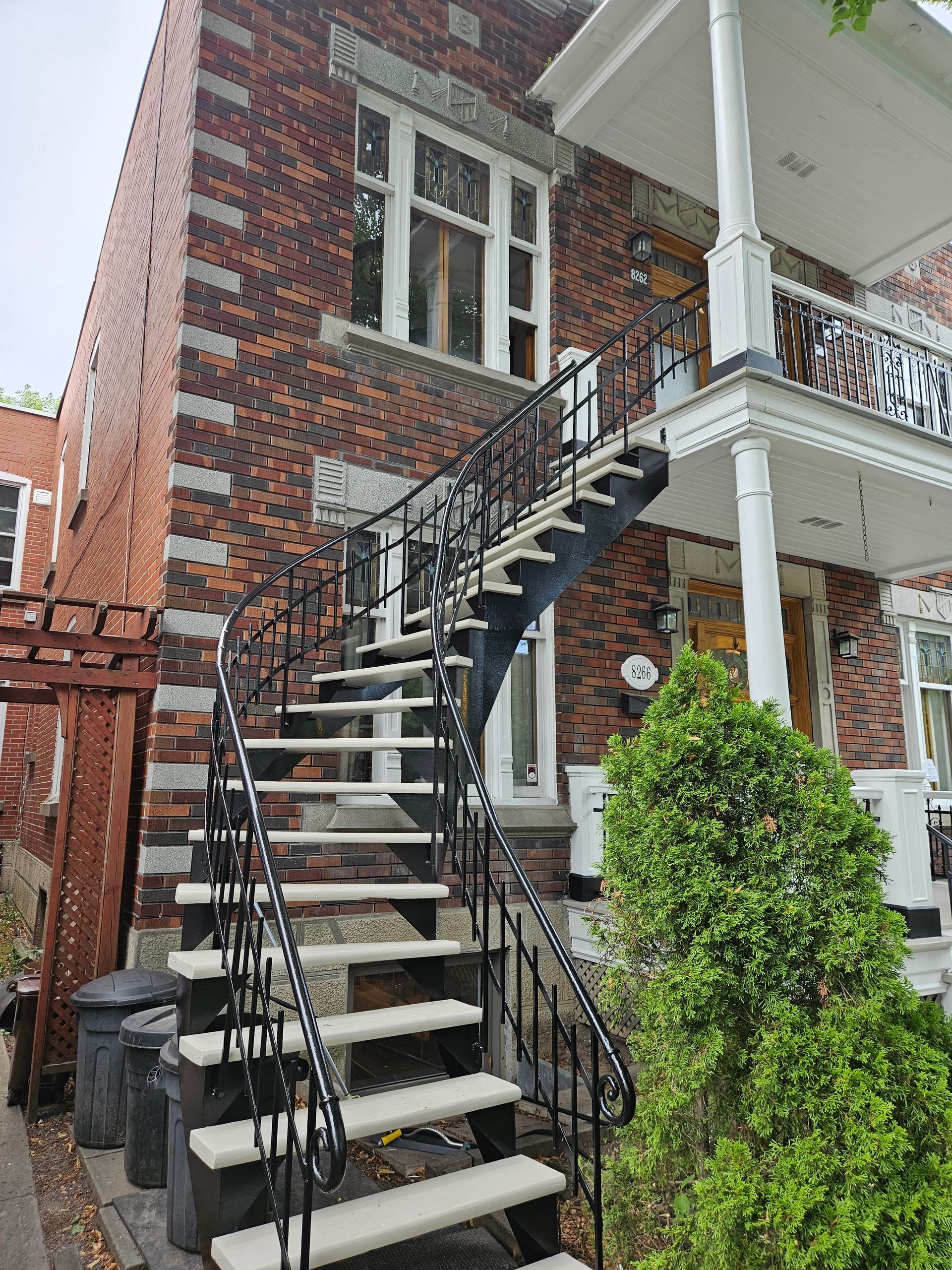 Bâtiment en briques avec escalier extérieur. Rambarde en fer forgé noir, marches grises, buisson vert.