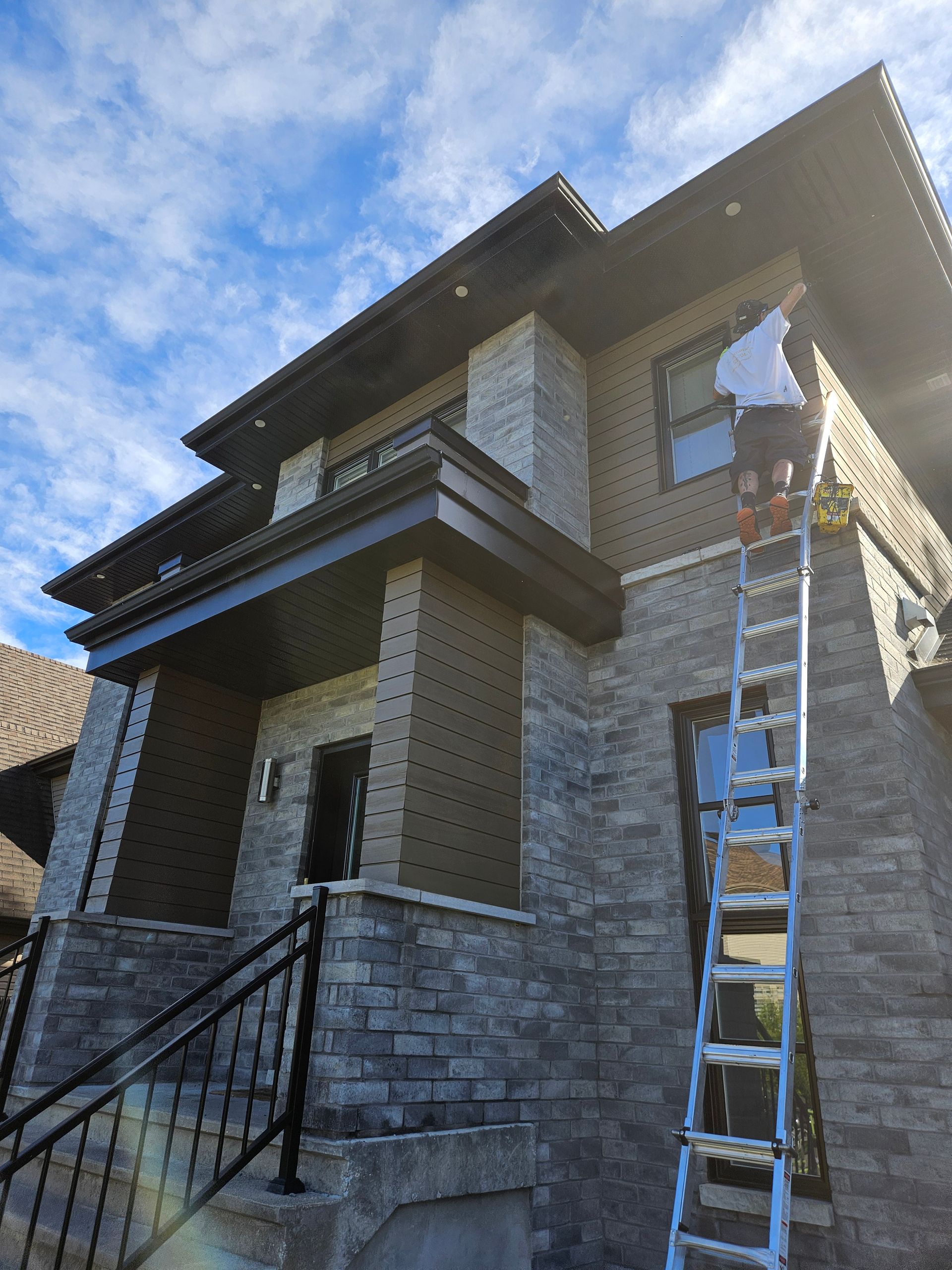 Une personne sur une échelle peint l'extérieur d'une maison à deux étages en pierre avec des boiseries brun foncé, sur fond de ciel partiellement nuageux.