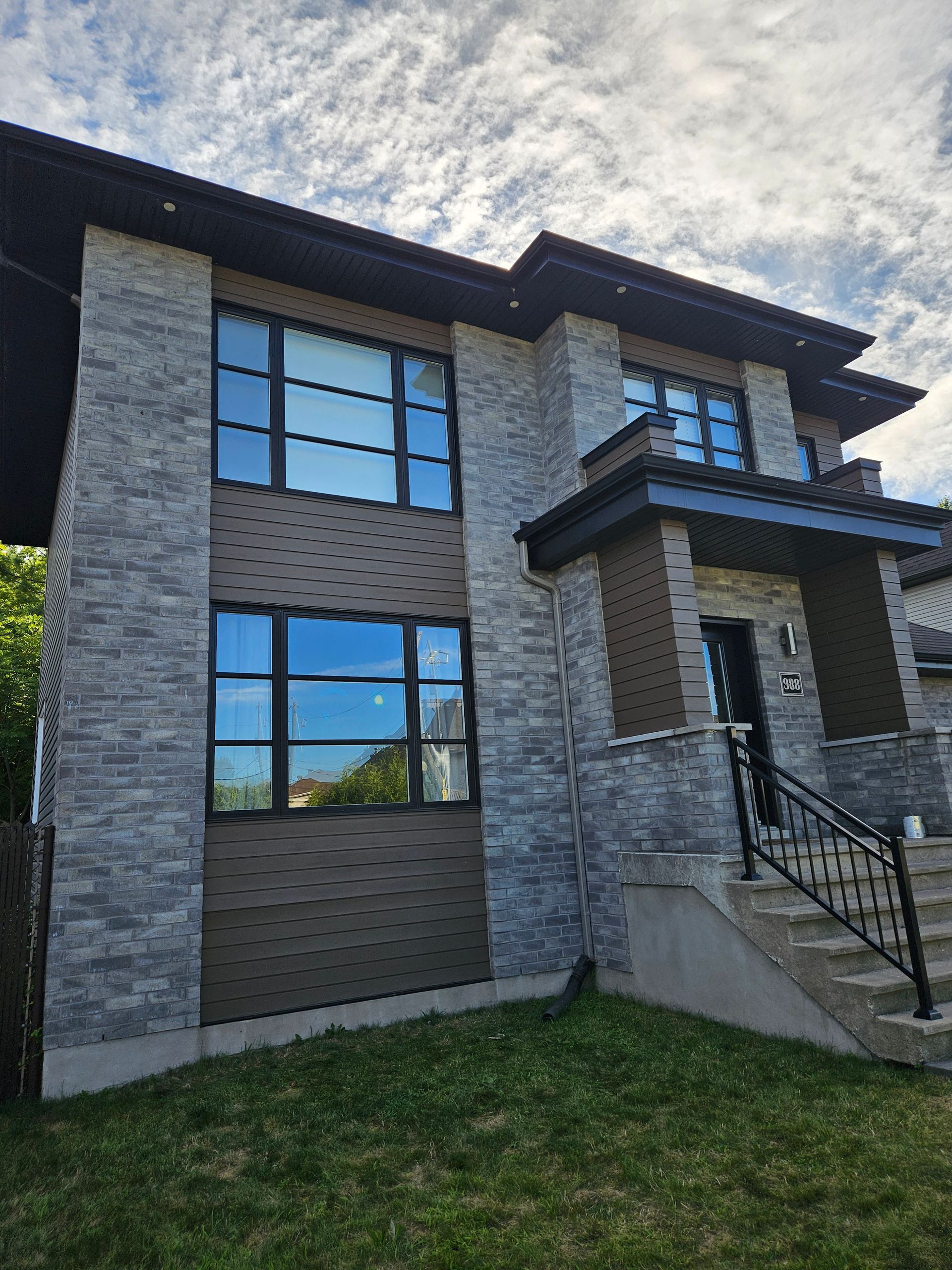 Two-story modern house with gray stone and brown accents, large windows, black roof, and small front yard.