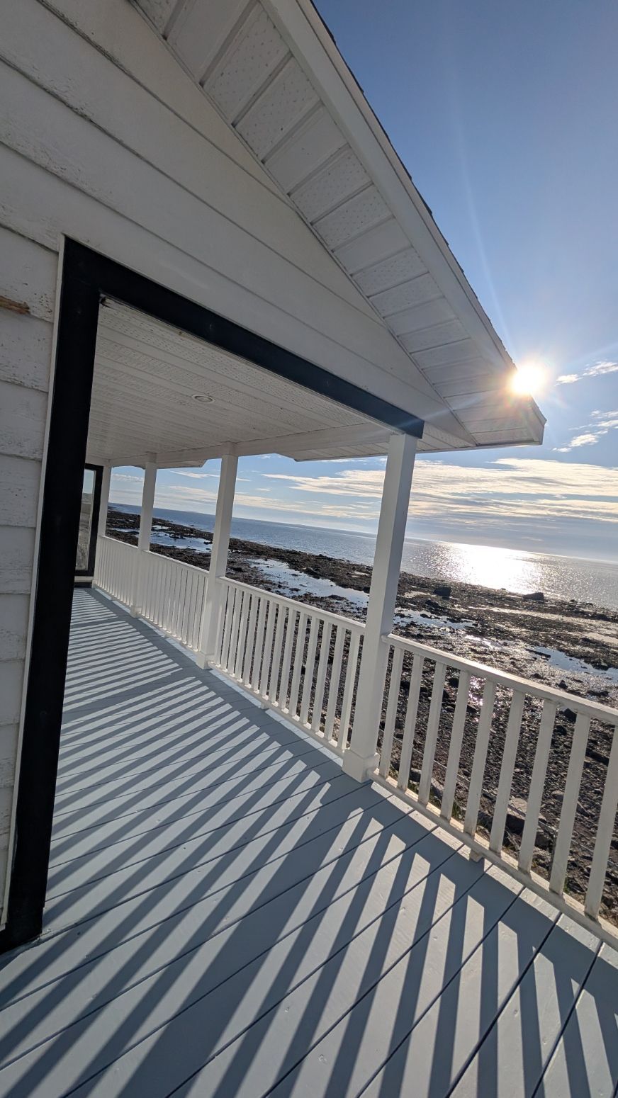 Véranda blanche donnant sur une mer ensoleillée. La lumière du soleil projette des ombres sur la terrasse bleu-gris.