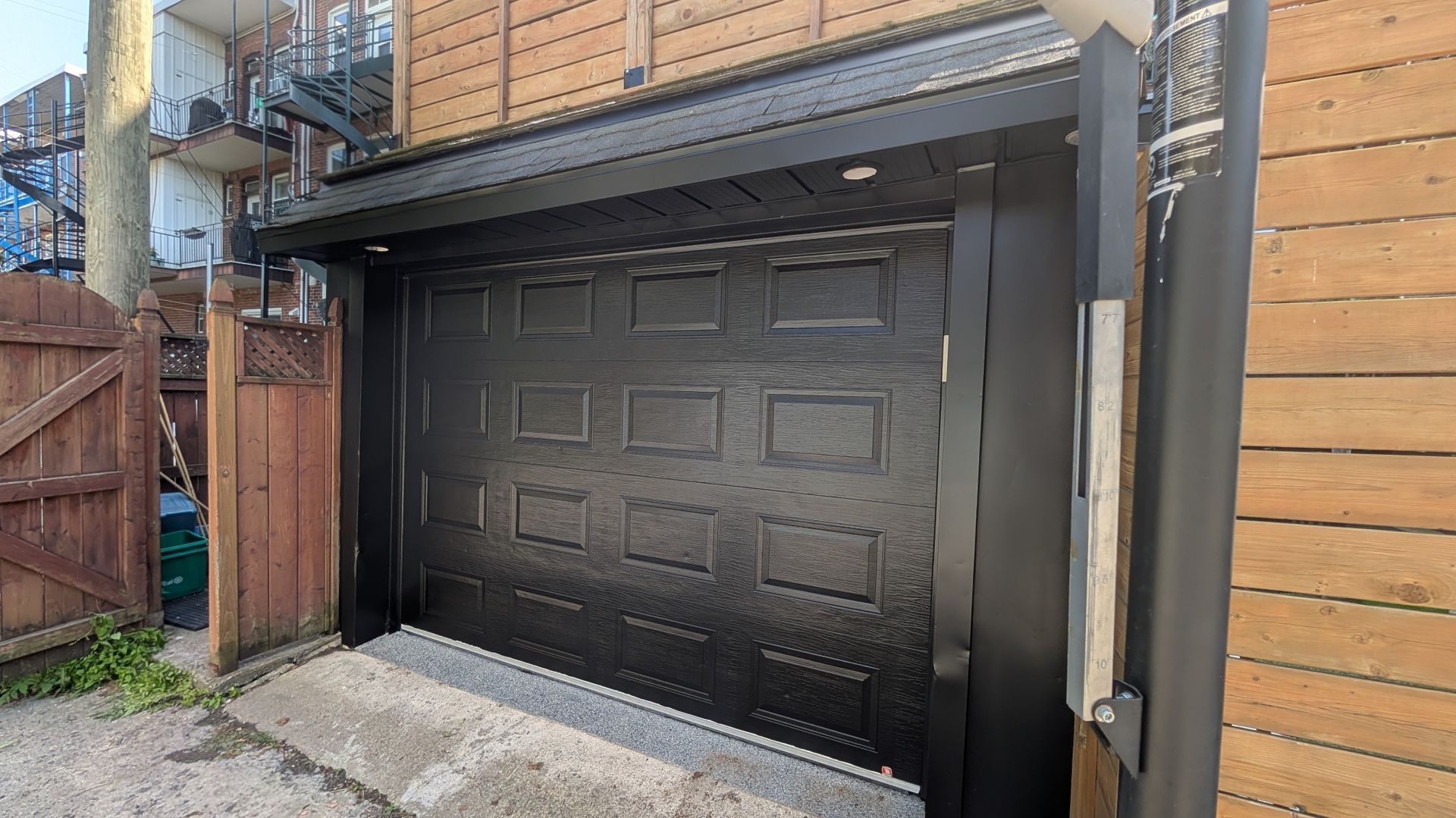 Porte de garage noire à panneaux texturés, à côté d'une clôture en bois et d'un bâtiment avec un escalier en métal.