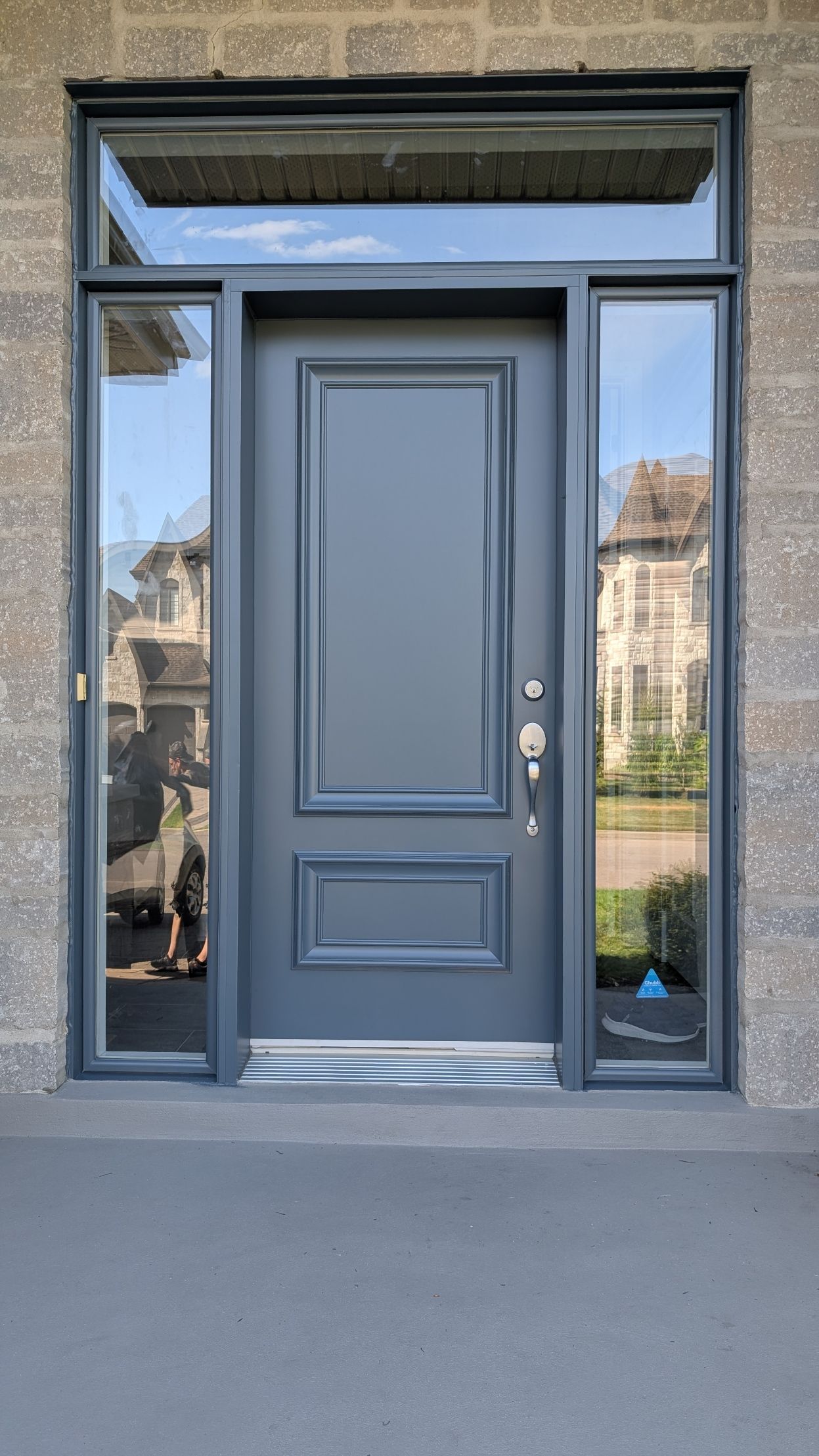 Porte d'entrée grise avec imposte et fenêtres latérales, intégrée à une façade en briques.