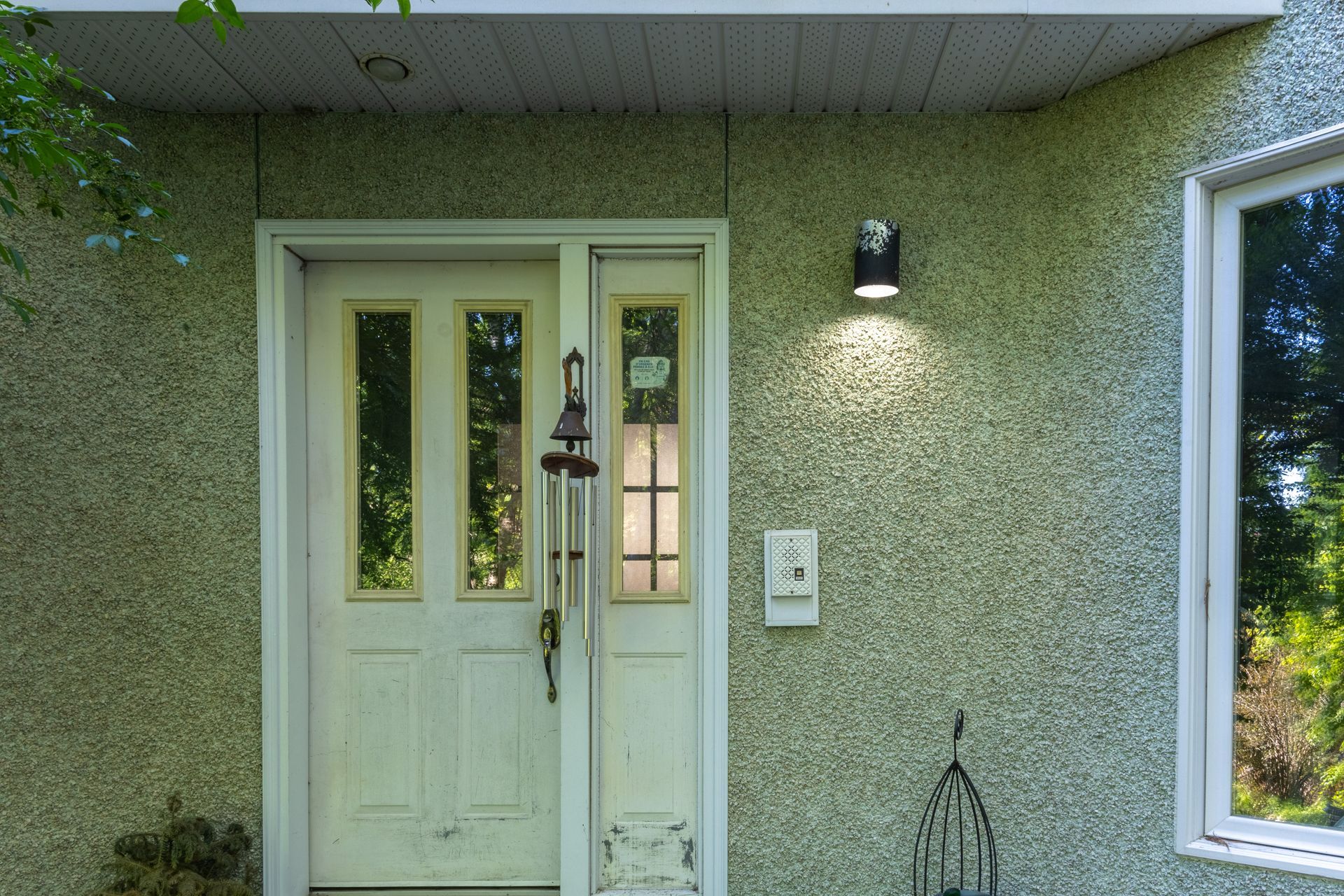 Porte d'entrée blanche avec panneaux de verre et façade en stuc vert texturé. Luminaire noir et cadre de fenêtre blanc.