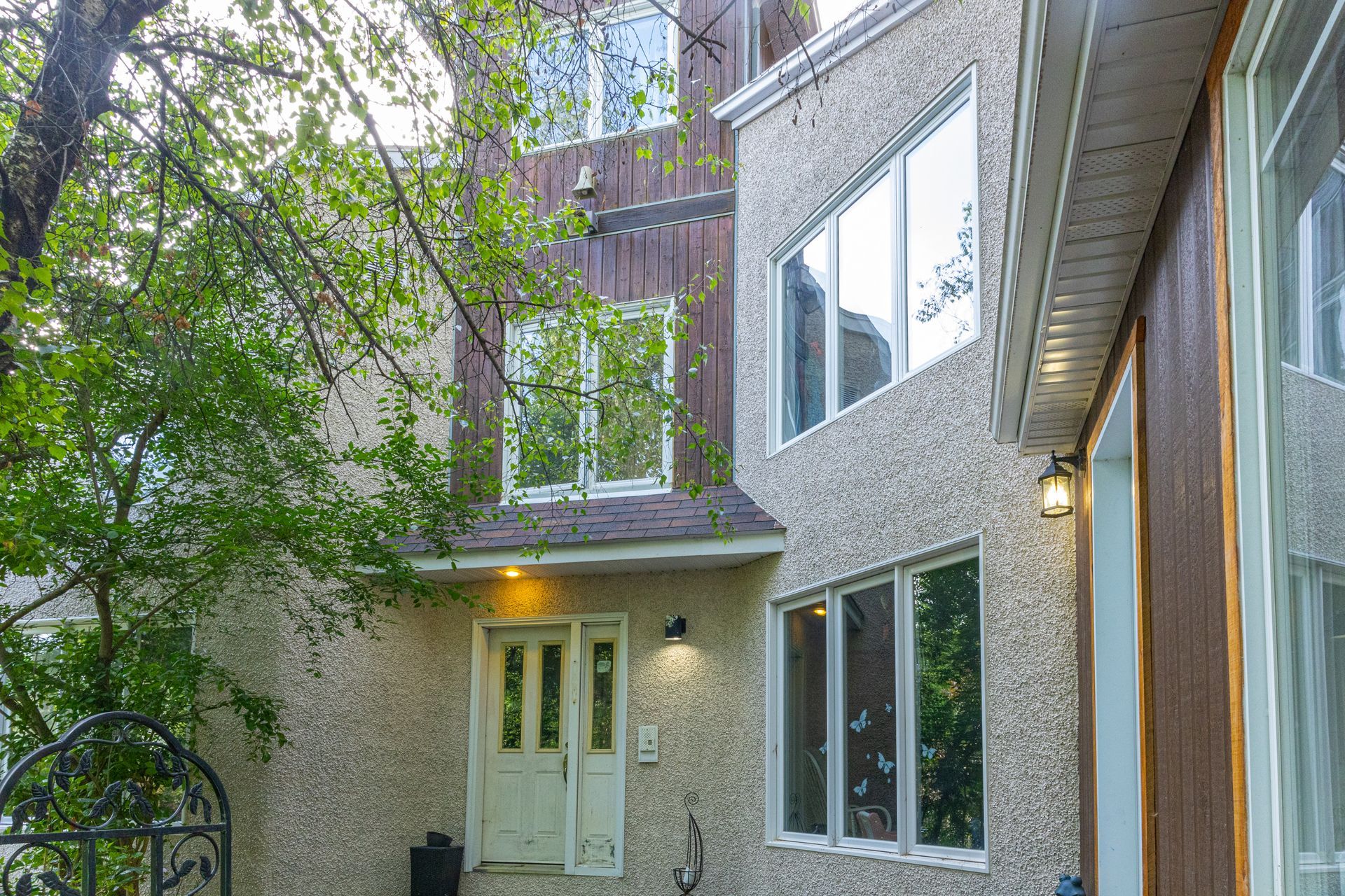 Vue extérieure d'une maison à plusieurs étages aux murs en stuc, avec fenêtres et porte entrouverte. Des arbres verts encadrent la vue.