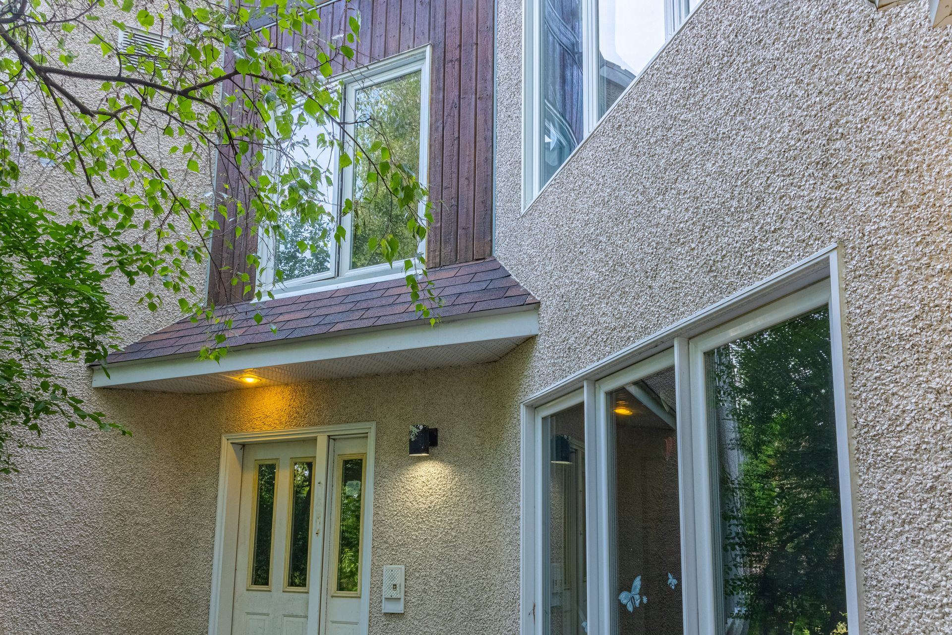 Façade en stuc beige et bois d'une maison à deux étages avec fenêtres à cadre blanc, une porte et un auvent marron.