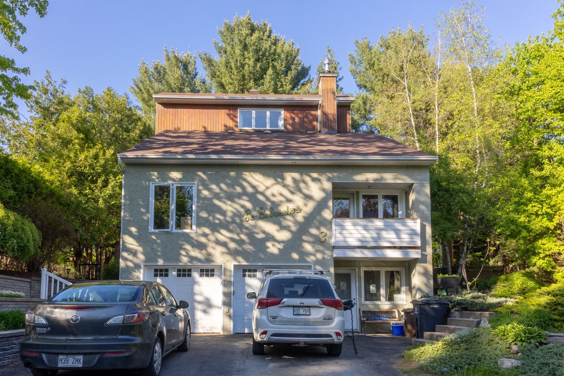 Maison à deux étages avec garage et deux voitures garées dans l'allée. Arbres et ciel bleu en arrière-plan.