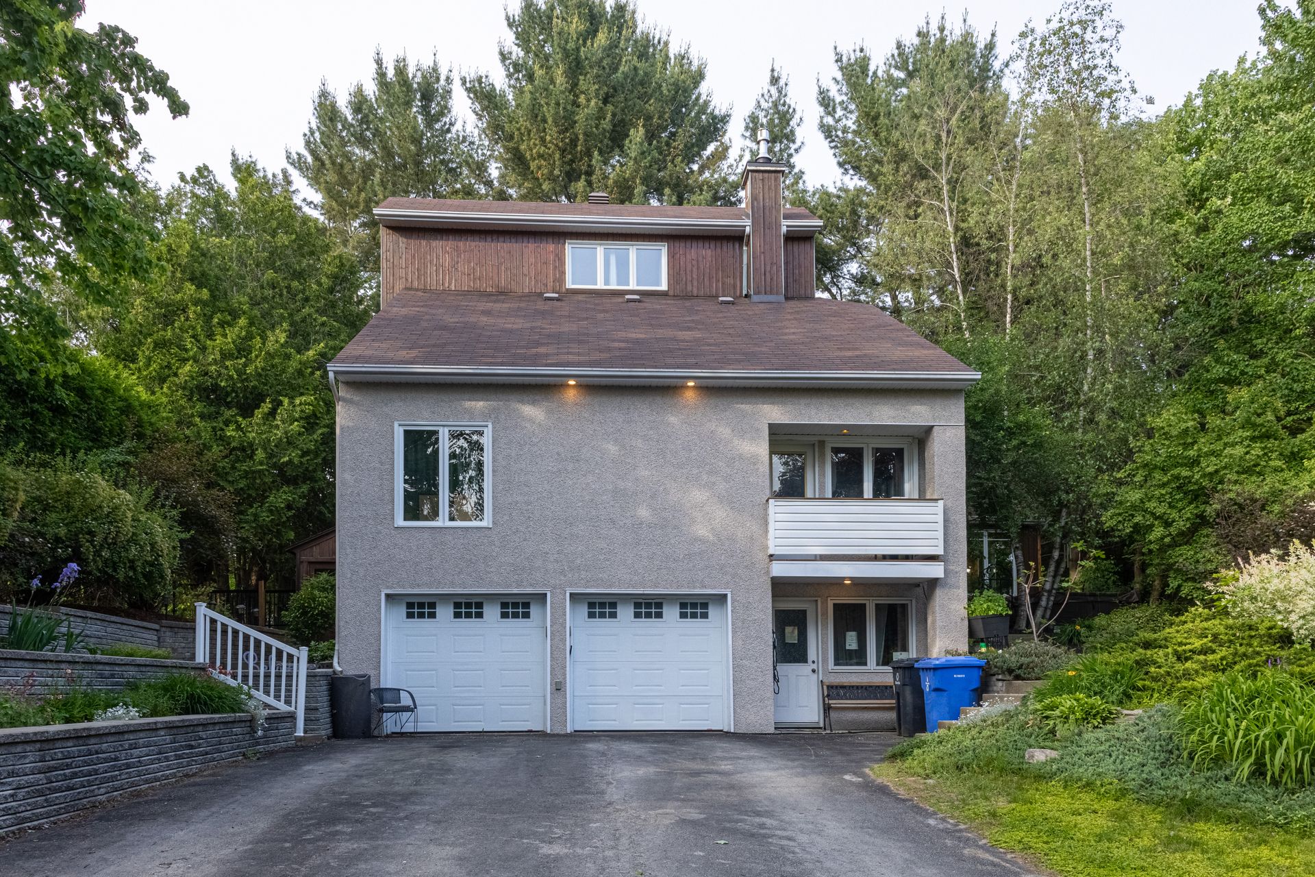 Maison à deux étages avec garage double, façade en stuc gris, toit marron, balcon, située dans une rue bordée d'arbres avec allée privée.
