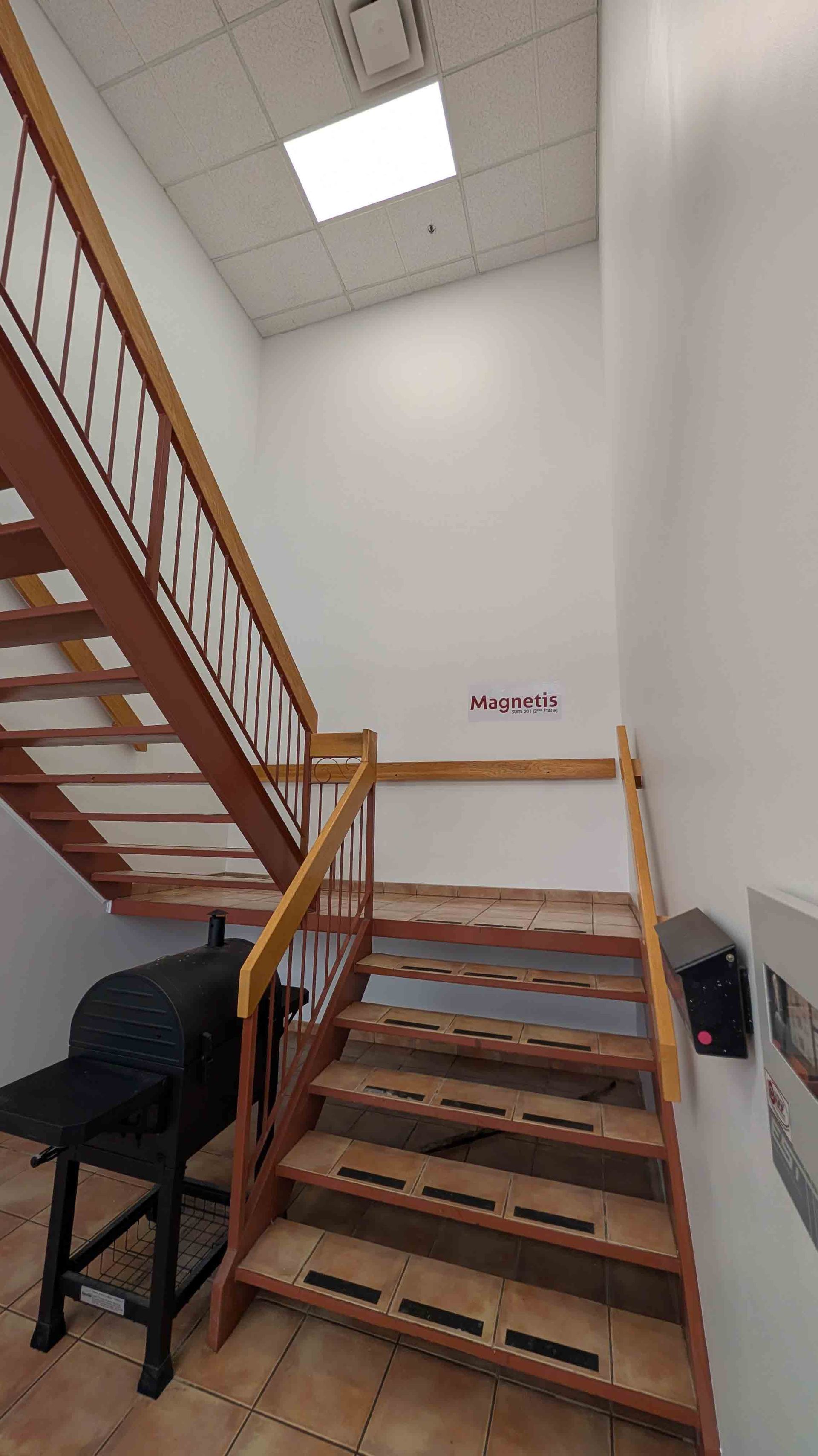 Cage d'escalier intérieure avec marches en bois, murs blancs et panneau lumineux au plafond.