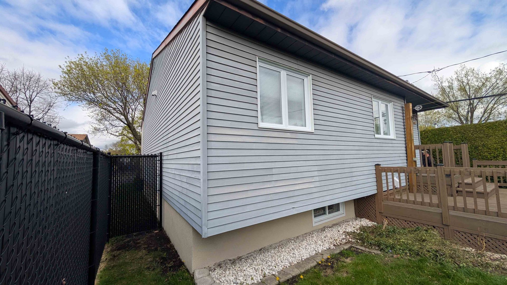Petite maison grise avec volets blancs, clôture noire et terrasse en bois dans la cour.