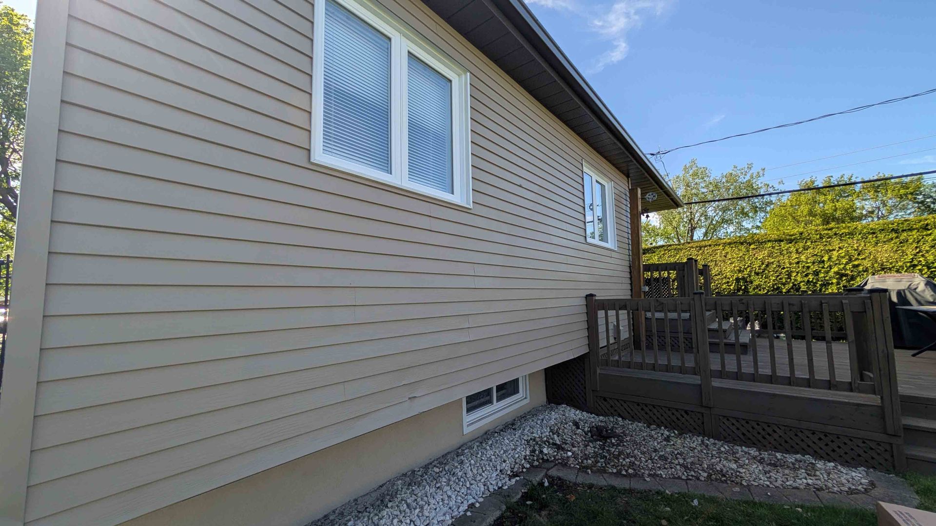 Vue latérale d'une maison beige aux fenêtres à encadrement blanc, aux fondations en gravier et à la clôture en bois, sous un ciel bleu.