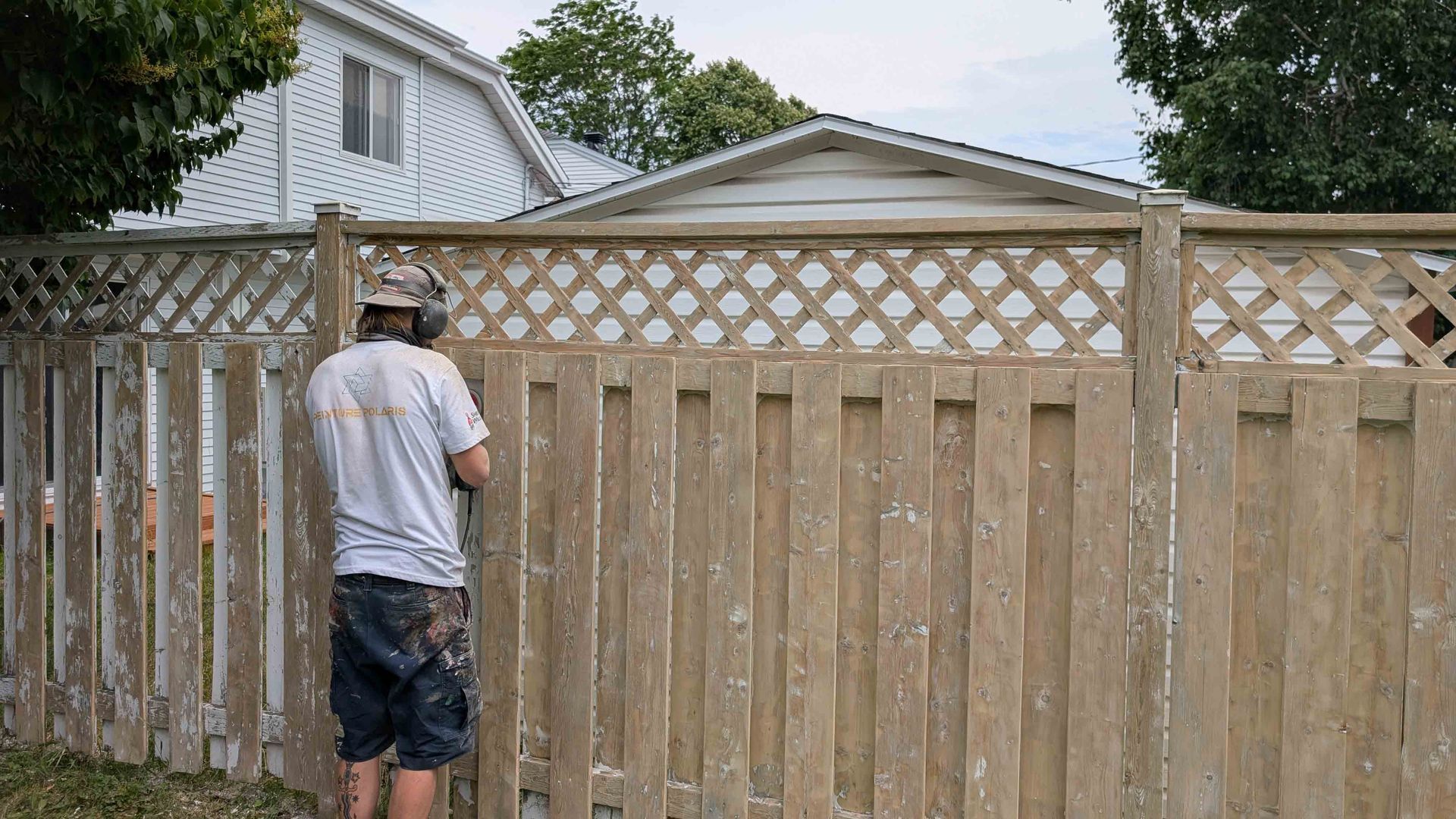 Une personne se tient près d'une clôture en bois, regardant à travers le treillis vers une maison et une cour.