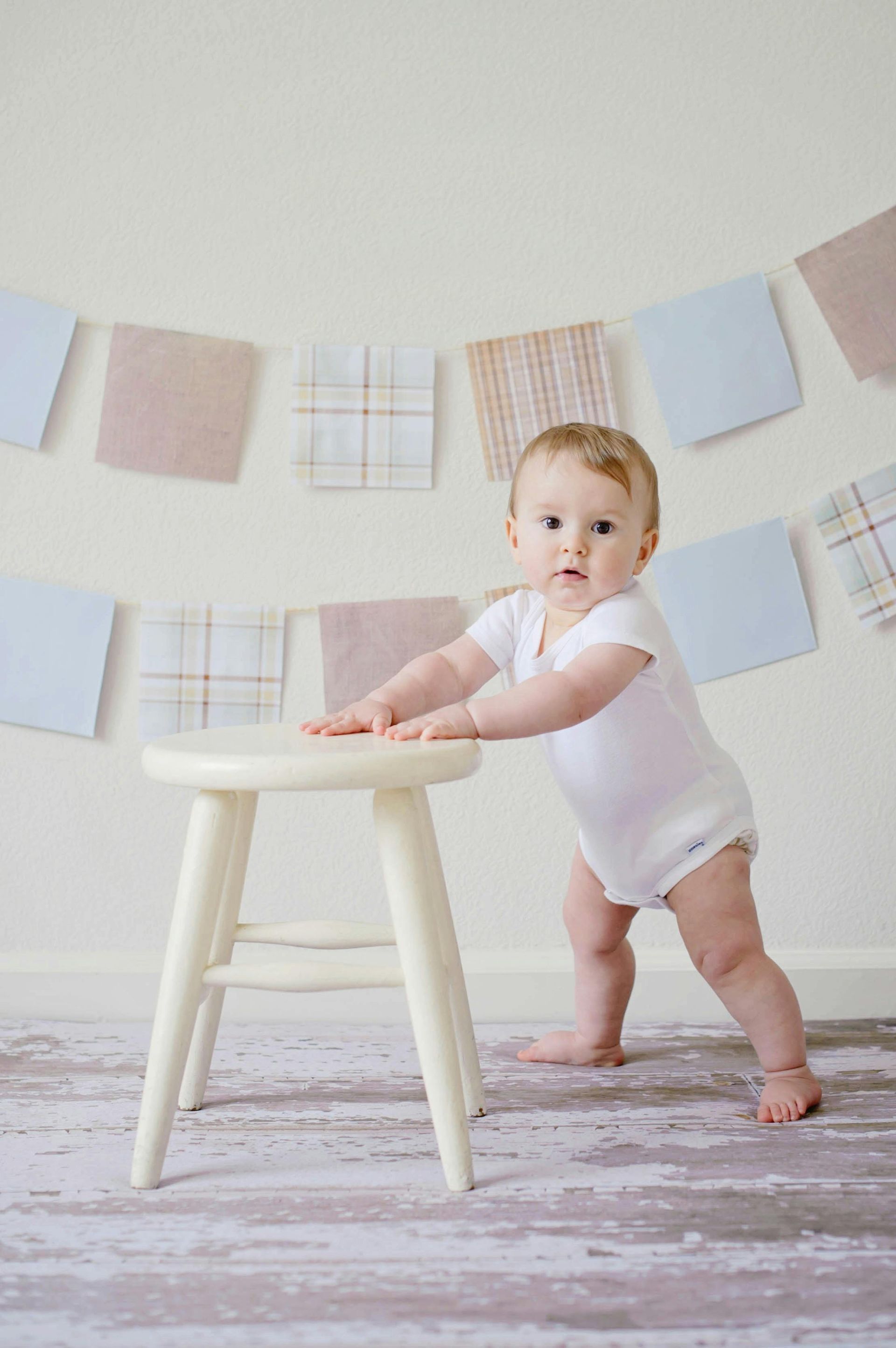 Um bebê está de pé ao lado de um banquinho branco.