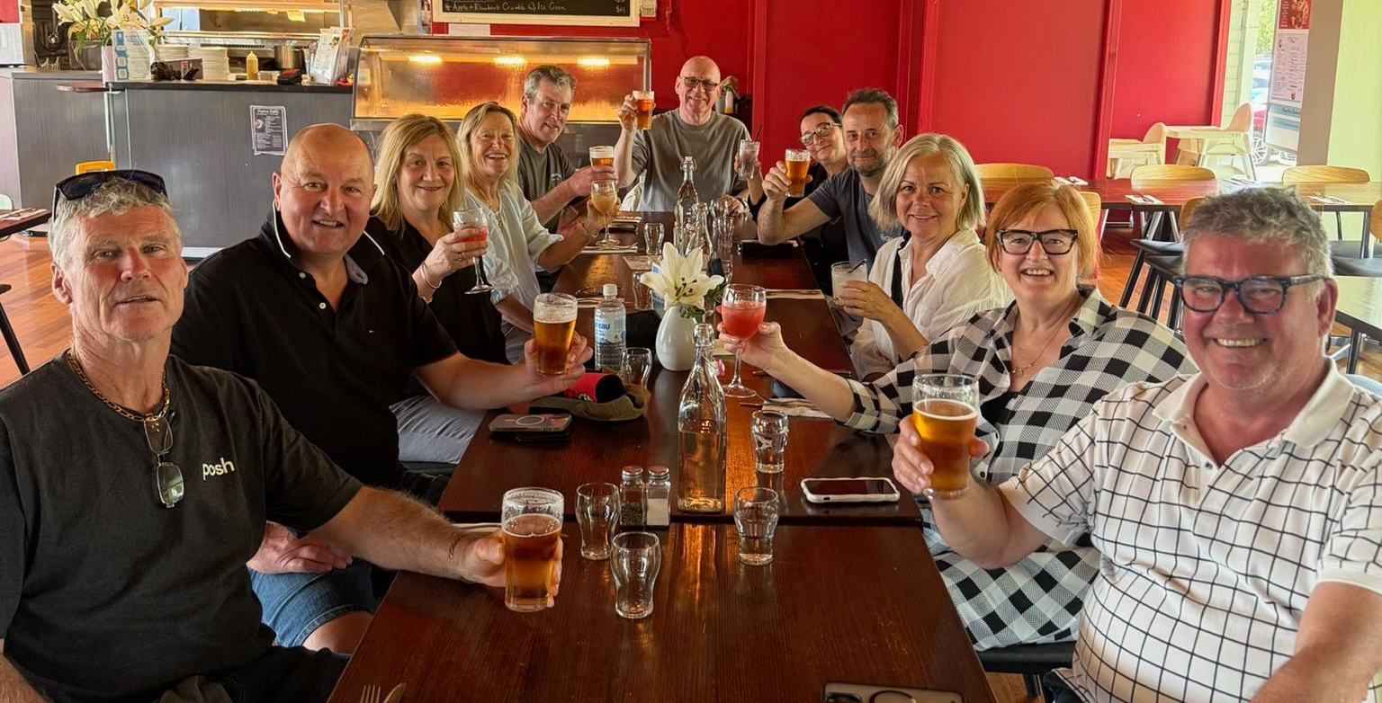 Group of people sitting at a table in restaurant drinking beer and toasting to the camera.