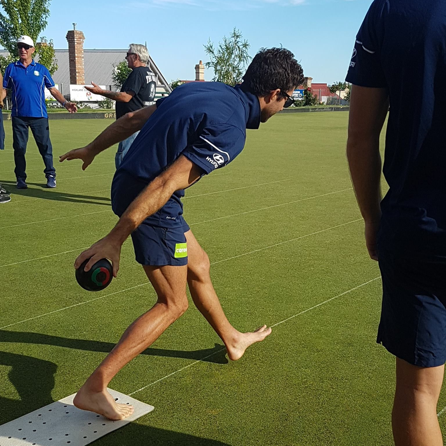 Young man on grass playing lawn bowls, wearing navy t-shirt and shorts.