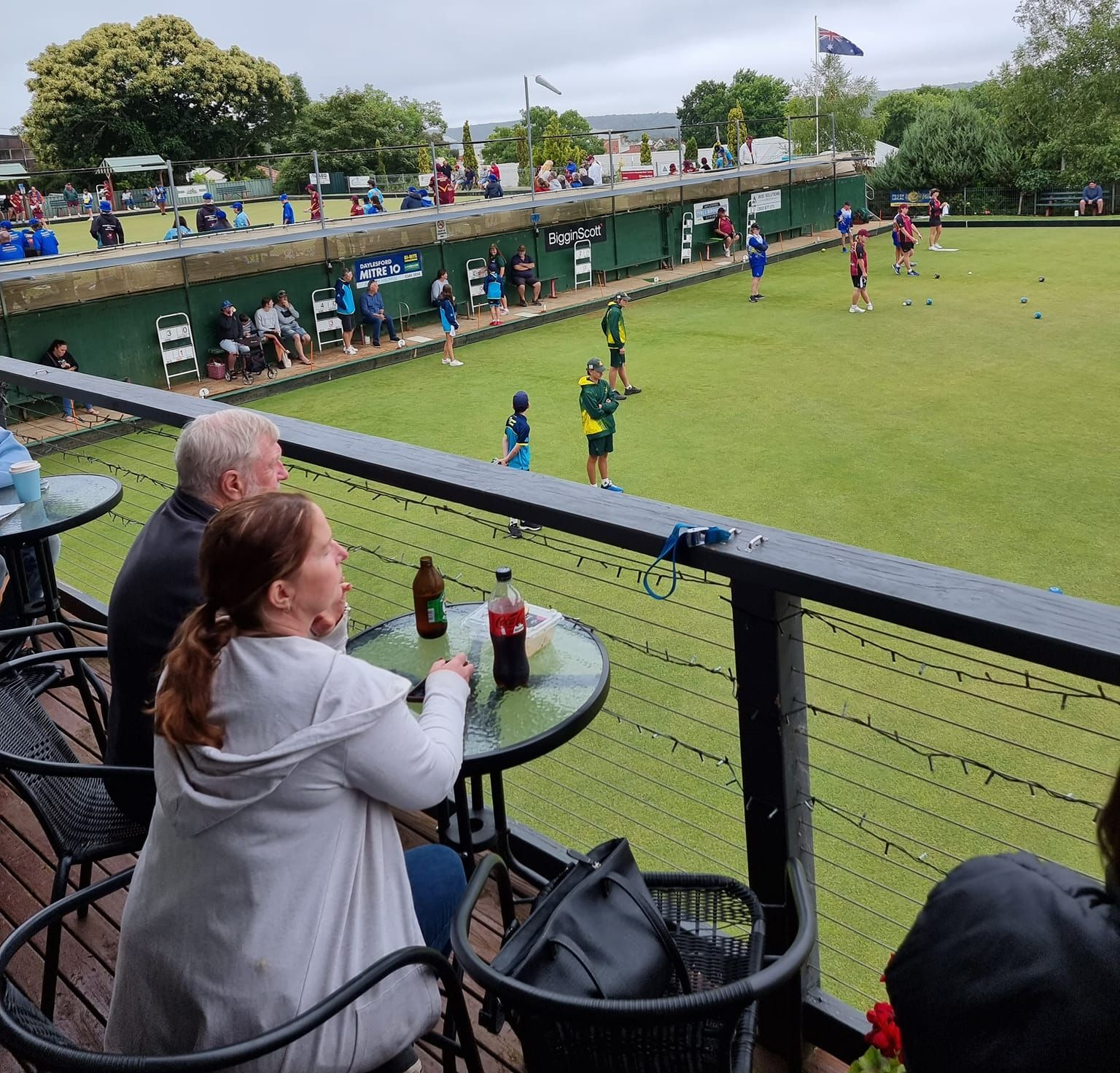 People watching lawn bowls from a balcony drinking at a small bistro table.