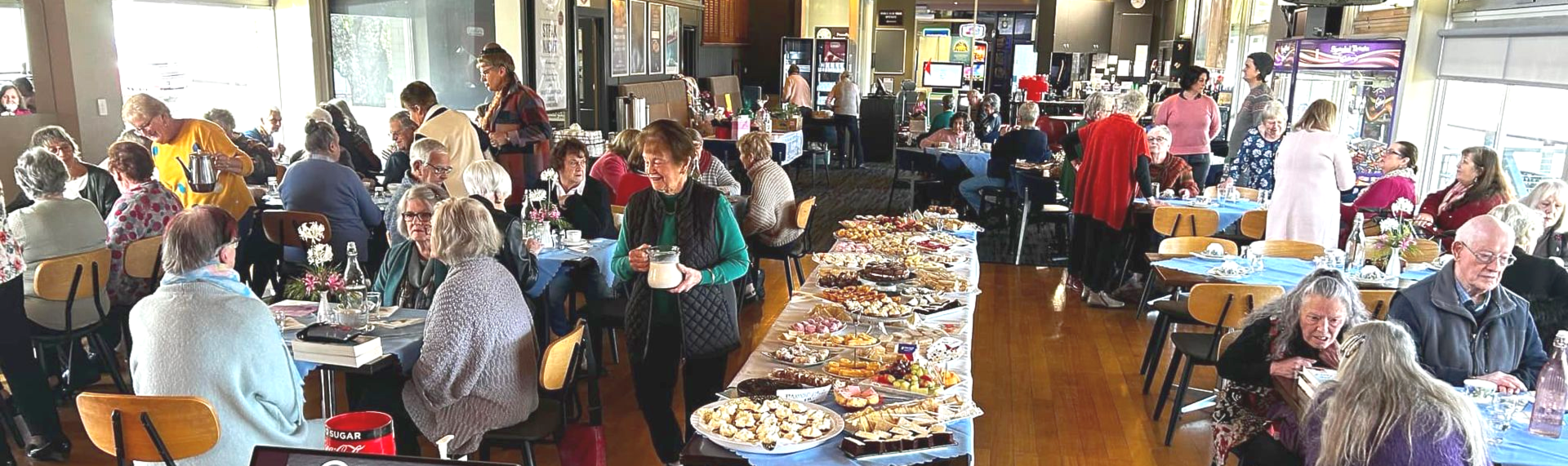 Image of people at a function. Some standing, some sitting. There is a long table with a buffet of food. 