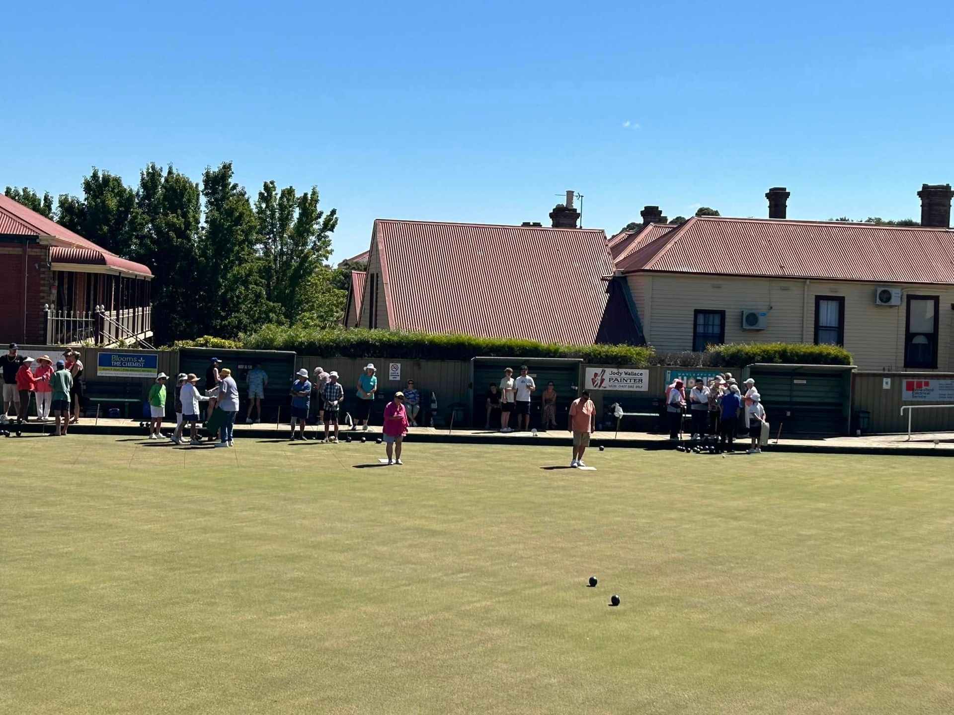 Bowling green with people and houses in background. There is a blue sky.