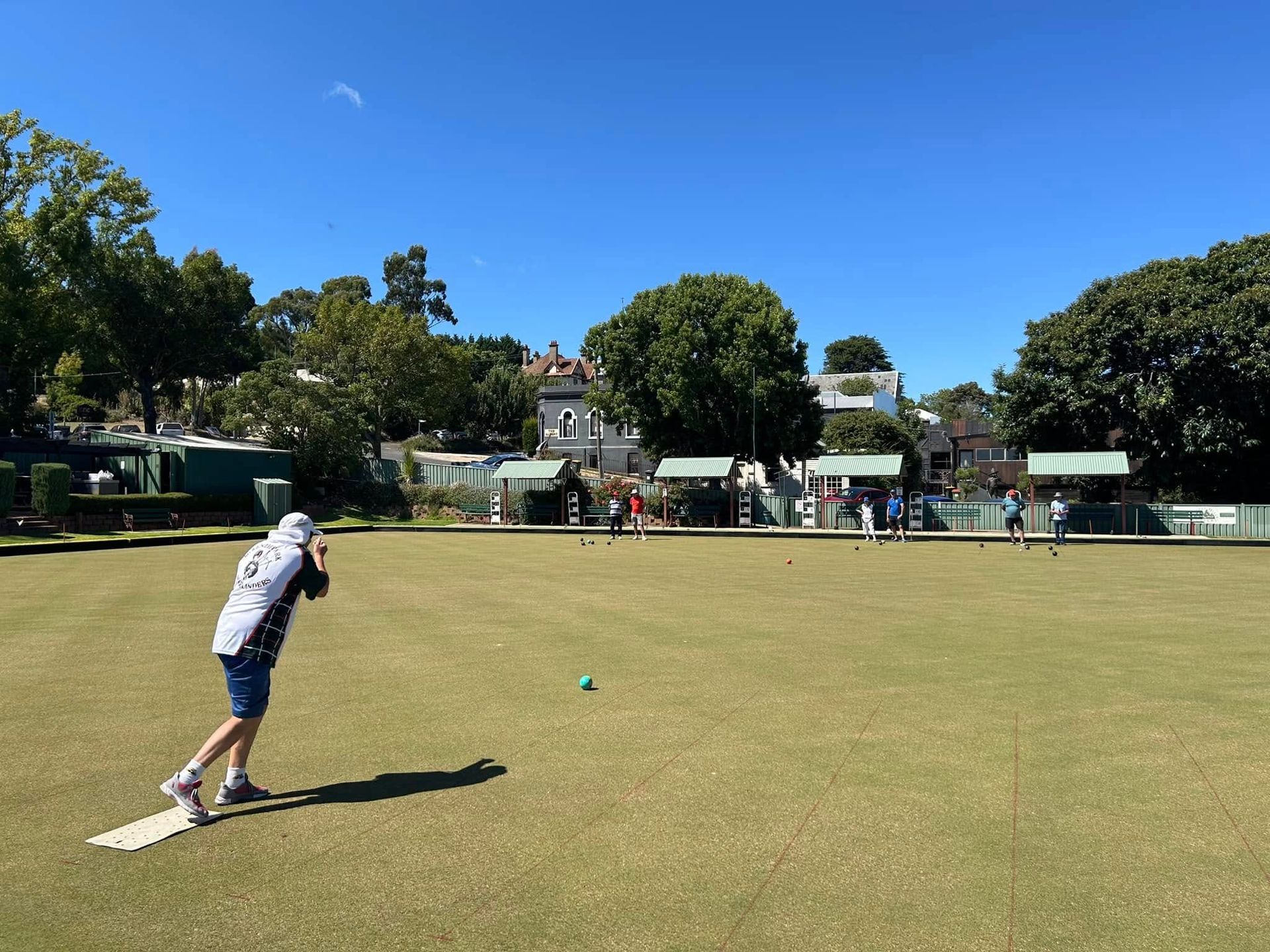 Bowling green with person in white shirt and hat bowling a green ball. There is a blue sky.