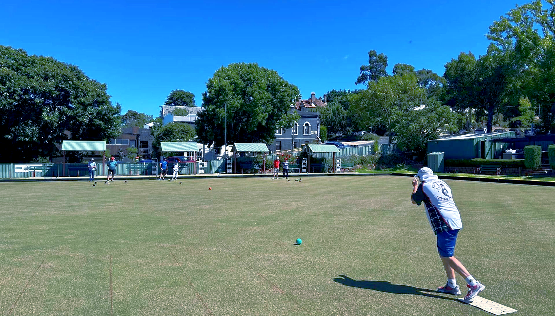 Person in white shirt on bowling green throwing a green ball. There is blue sky.