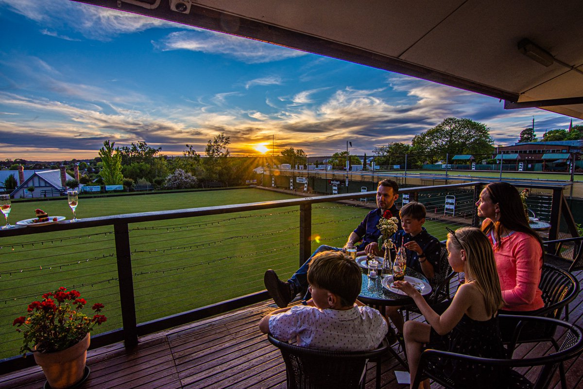 Young family sitting on veranda of lawn bowls venue on balcony at sunset.