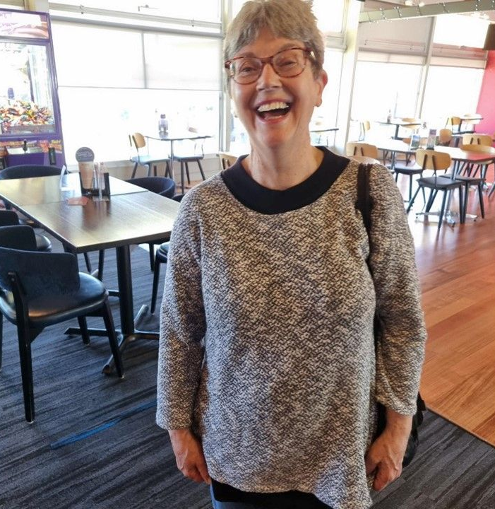 Lady smiling showing teeth. She has glasses, grey short hair and is wearing a grey jumper. She is standing in a restaurant with empty tables and chairs behind her.