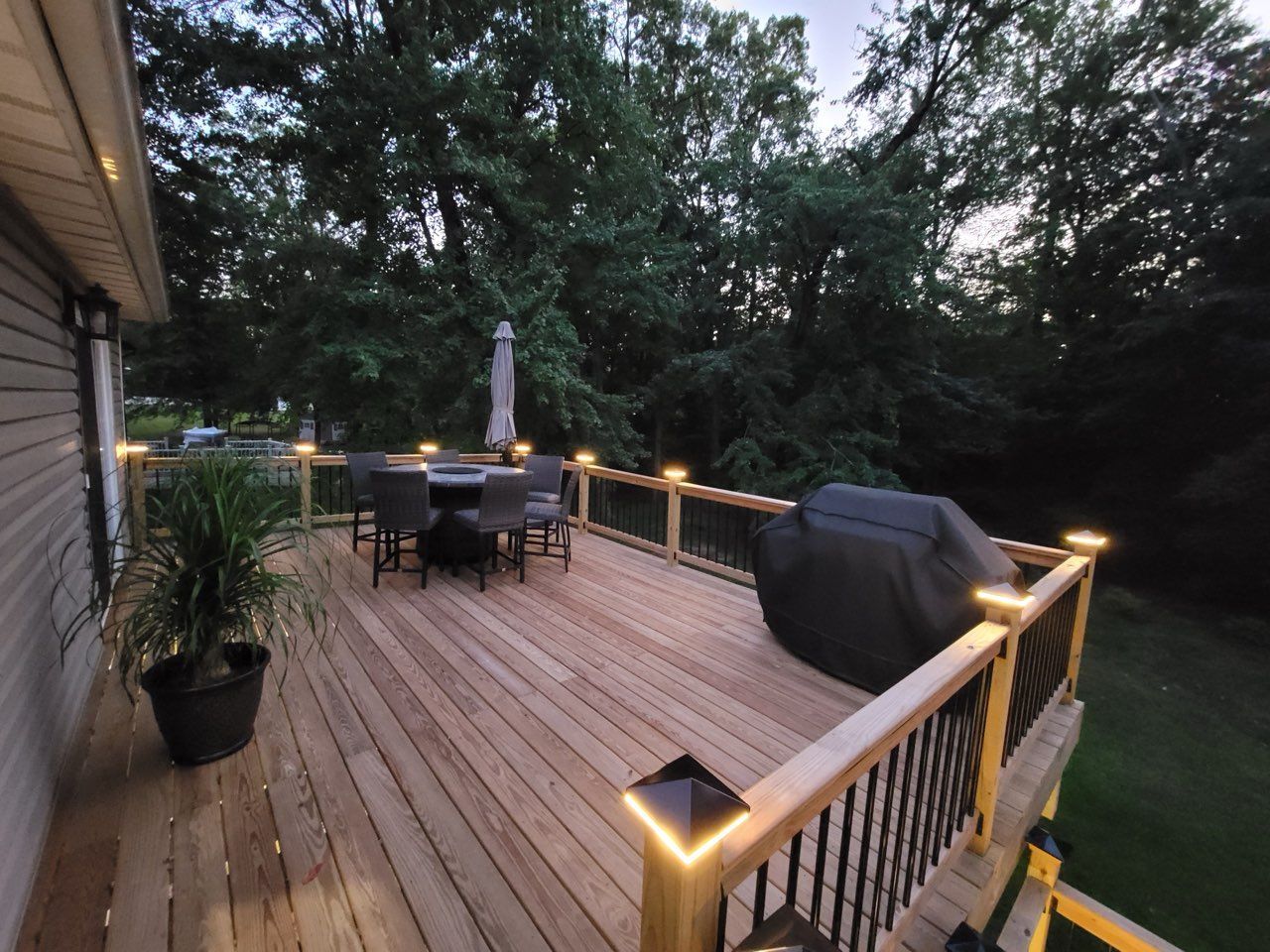 Wooden deck with railing lights, patio table and grill, overlooking trees at dusk