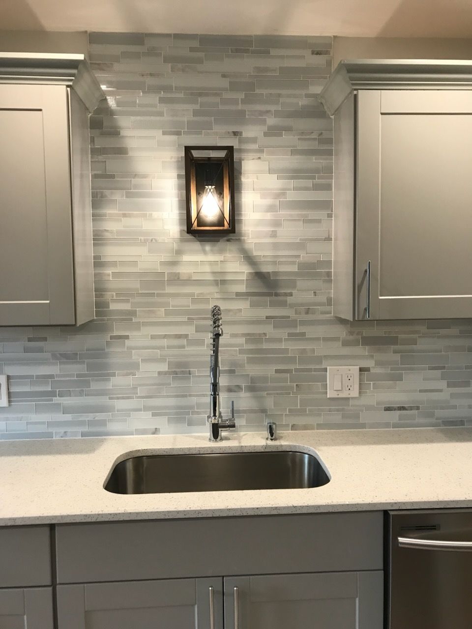 Modern kitchen sink with gray cabinets, white countertop, and gray tile backsplash with wall light