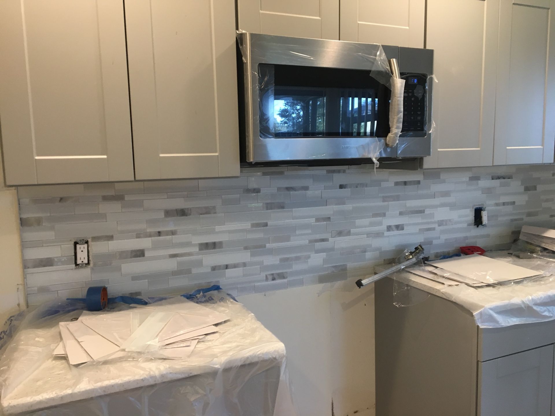 Kitchen under renovation with white cabinets, mosaic backsplash, and a microwave wrapped in protective plastic.