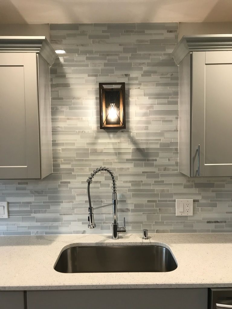 Modern kitchen sink with gooseneck faucet, white countertops, and gray tile backsplash under a wall sconce