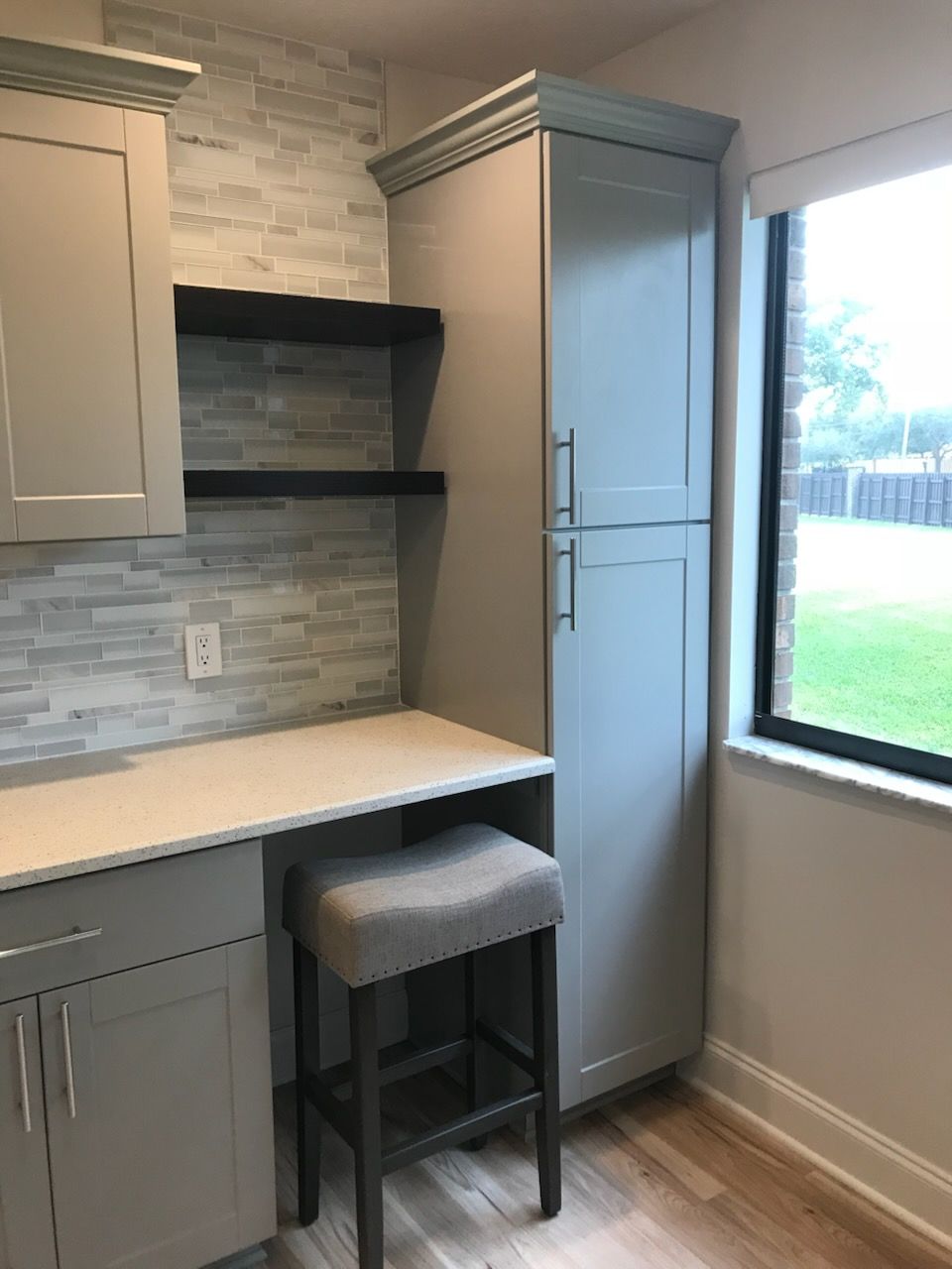 Bright kitchen nook with gray cabinets, stone backsplash, corner desk, stool, and a window overlooking a grassy yard