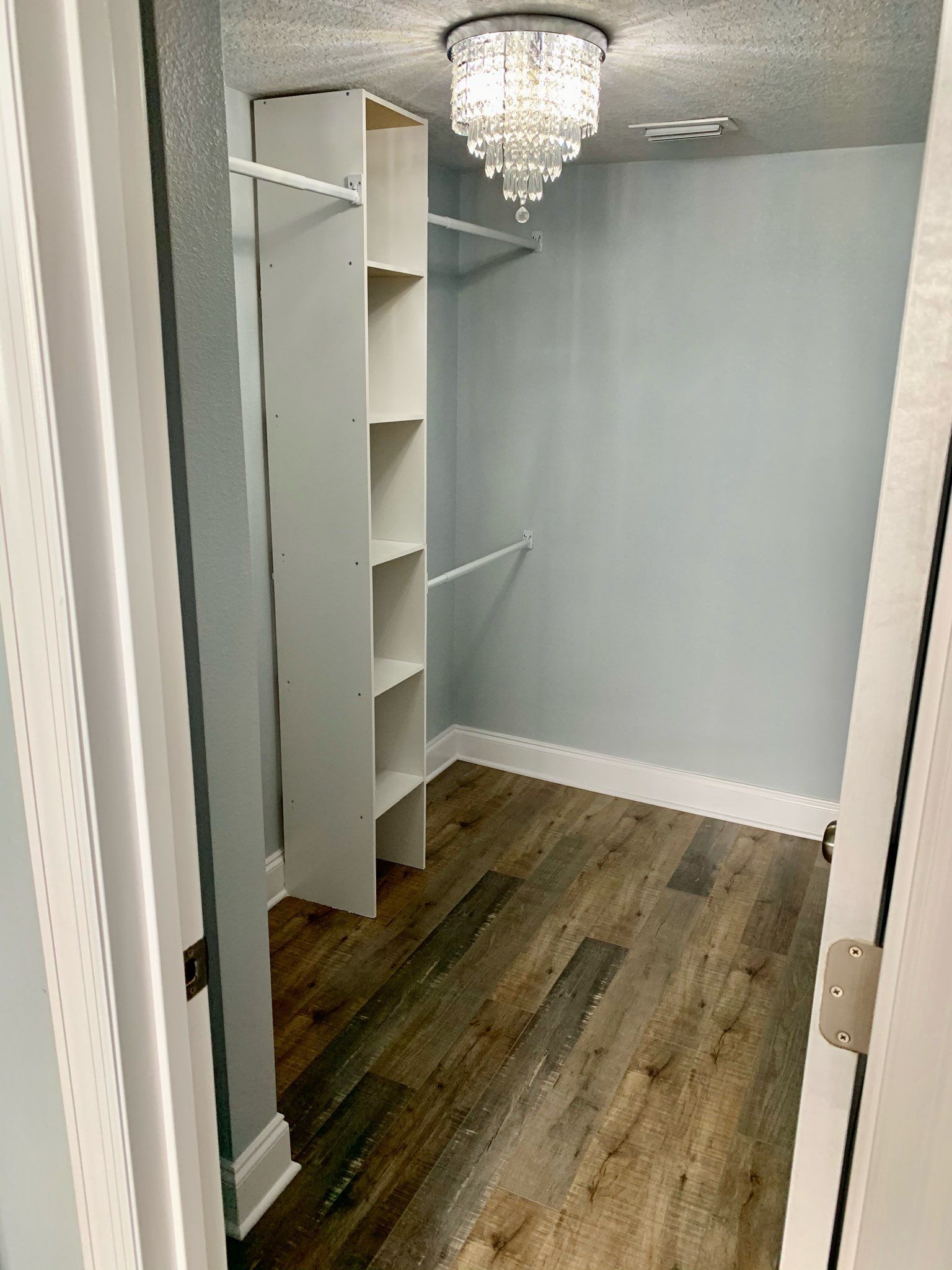 Small walk-in closet with white shelves, gray walls, wood floor, and a chandelier overhead.