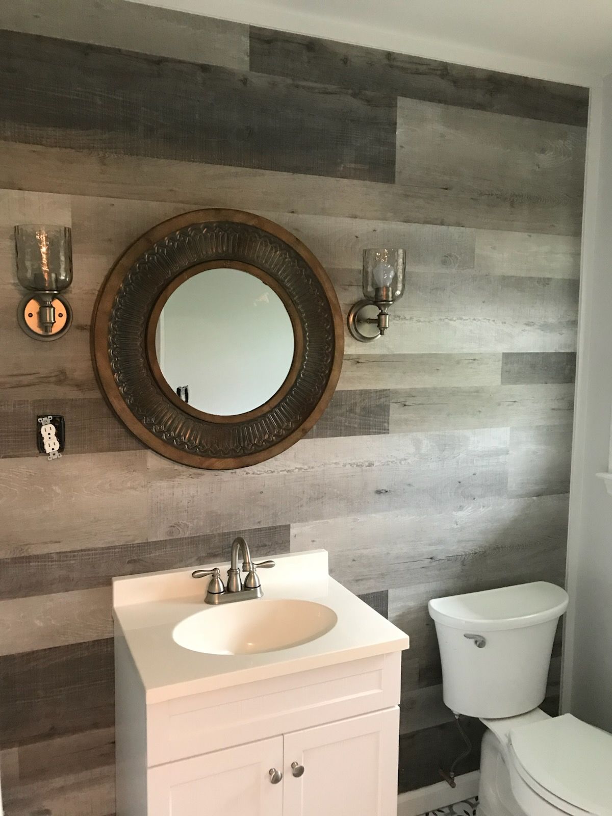 Small bathroom with gray wood panel walls, round mirror, white vanity, and toilet.