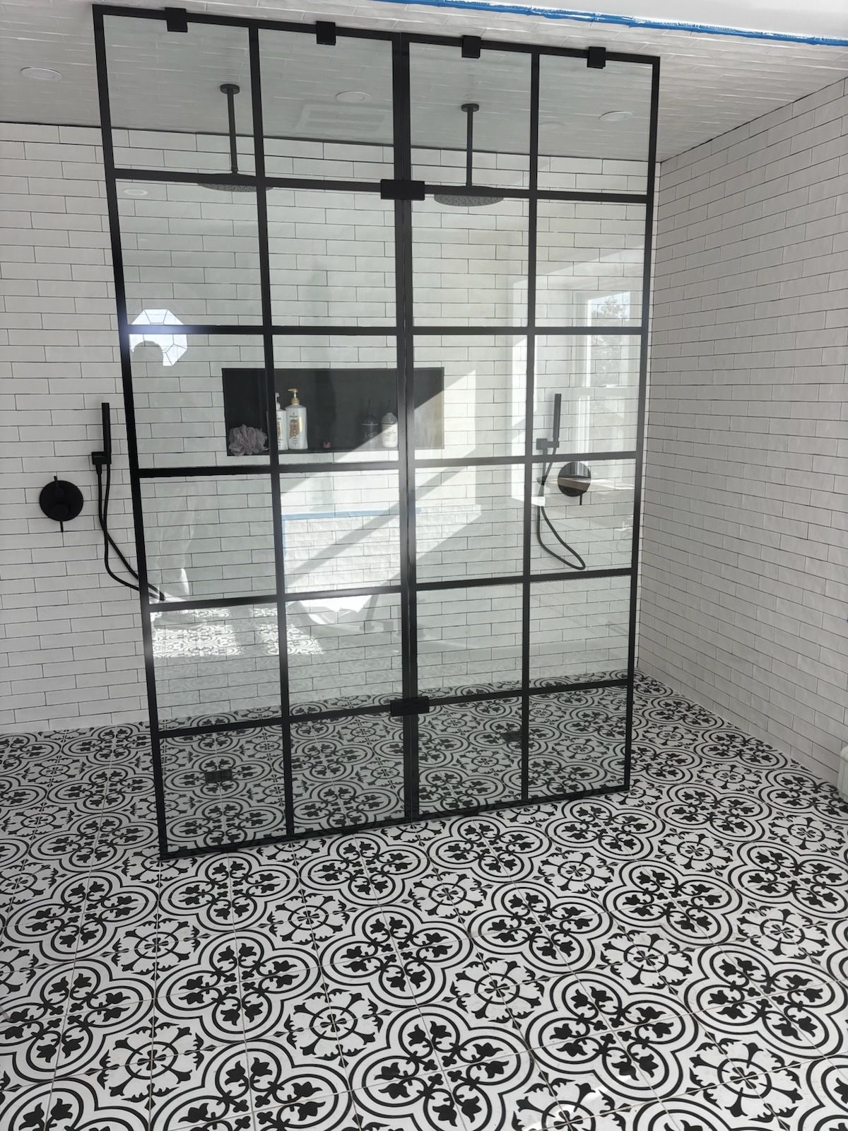 Black metal-framed glass shower enclosure in a tiled bathroom with patterned black-and-white floor.