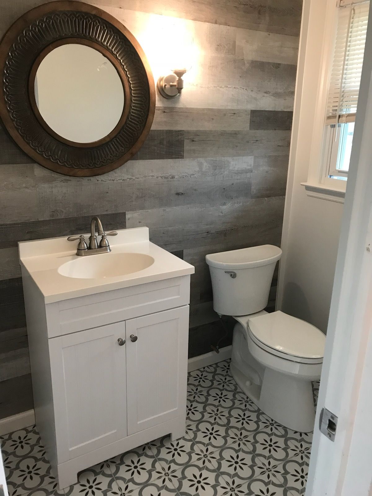 Small bathroom with white vanity, toilet, round mirror, and patterned tile floor.