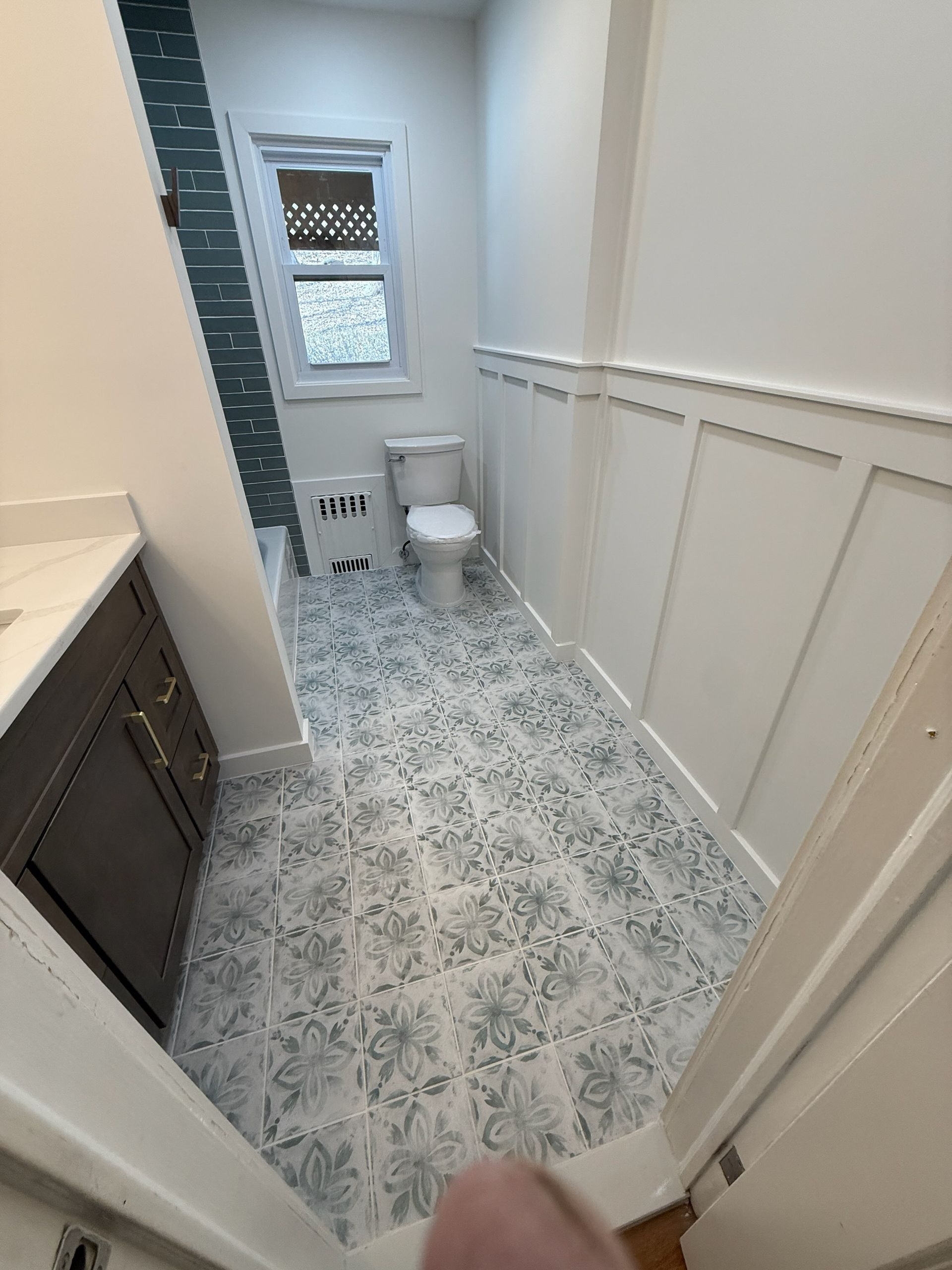 Narrow bathroom with white wainscoting, patterned tile floor, vanity, toilet, and window.