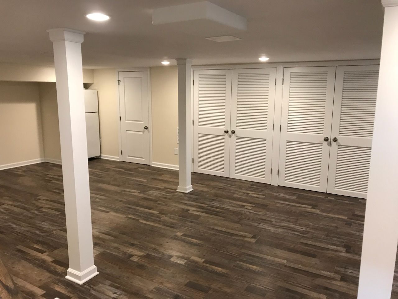 Empty finished basement room with dark wood floor, white columns, and louvered closet doors