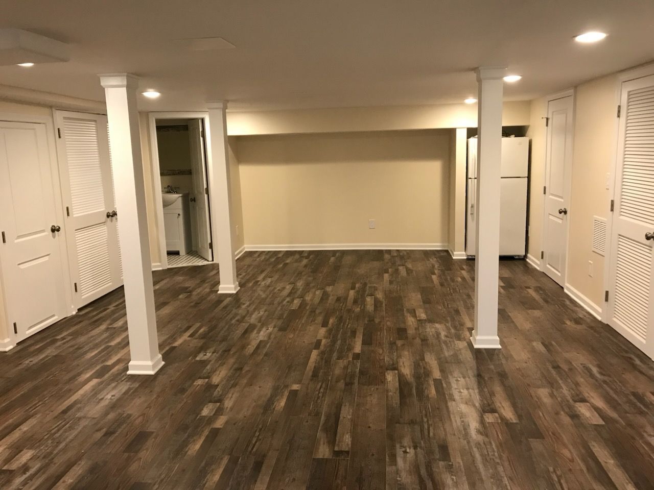 Empty finished basement room with dark wood flooring, white columns, recessed lights, and closet doors.