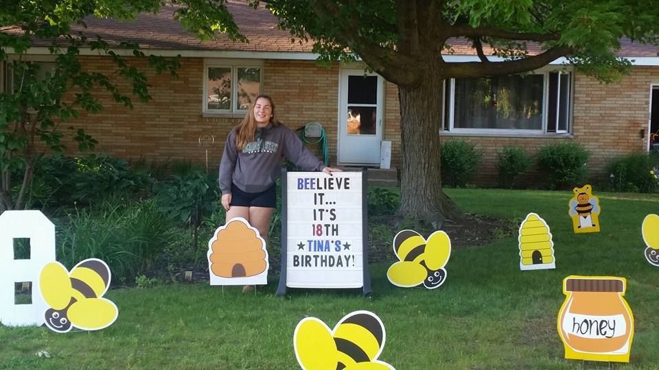 A woman is standing in front of a house with a sign that says believe it.  Bee themed yard card.