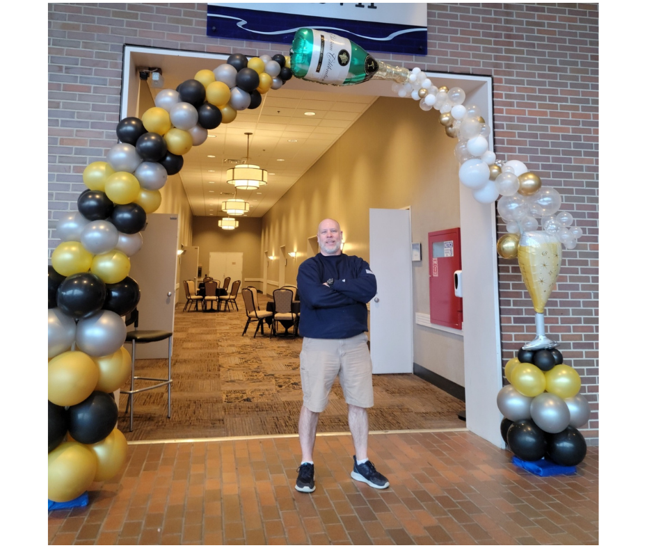 A man is standing in front of a balloon arch with a bottle of champagne coming out of it.