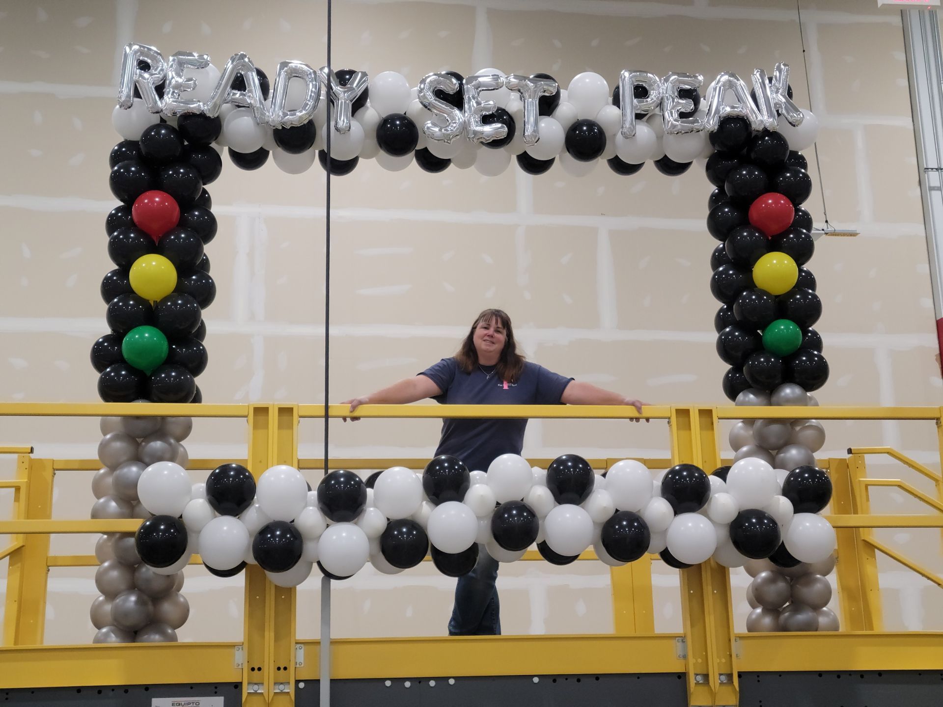 A woman stands in front of a balloon arch that says ready set peak at Amazon.  Racecar theme.
